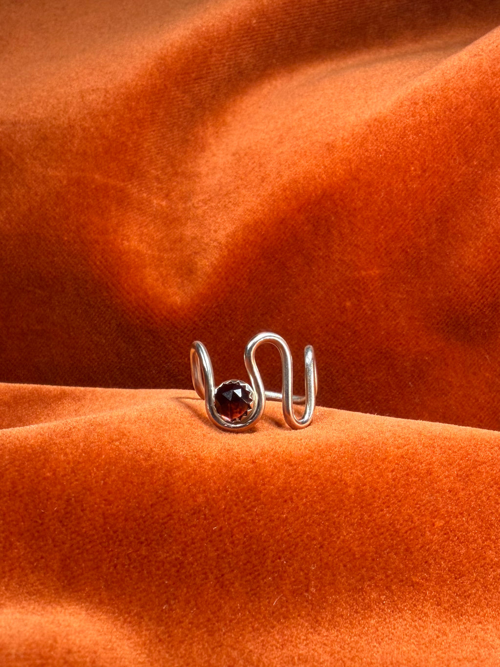 Close up image of silver ring with a asymmetrical band. A red rose cut garnet stone is set in a serrate bezel floating between the wavy band. 