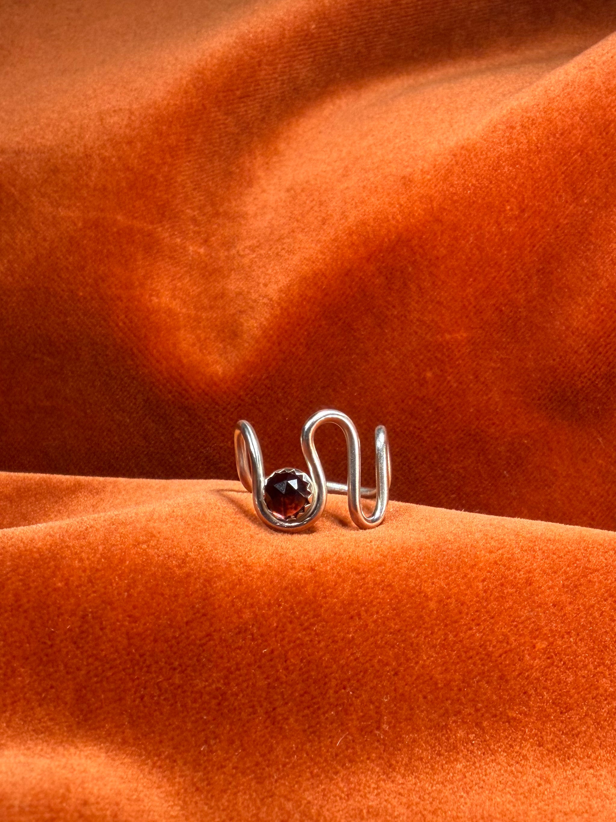 Close up image of silver ring with a asymmetrical band. A red rose cut garnet stone is set in a serrate bezel floating between the wavy band. 