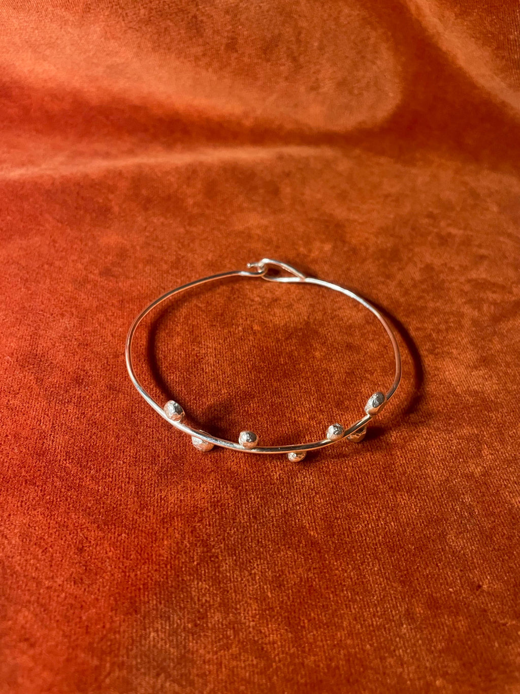 Top down view of a silver bangle with a hook clasp and seven small orbs on the top of the band. Stage on an orange velvet background.