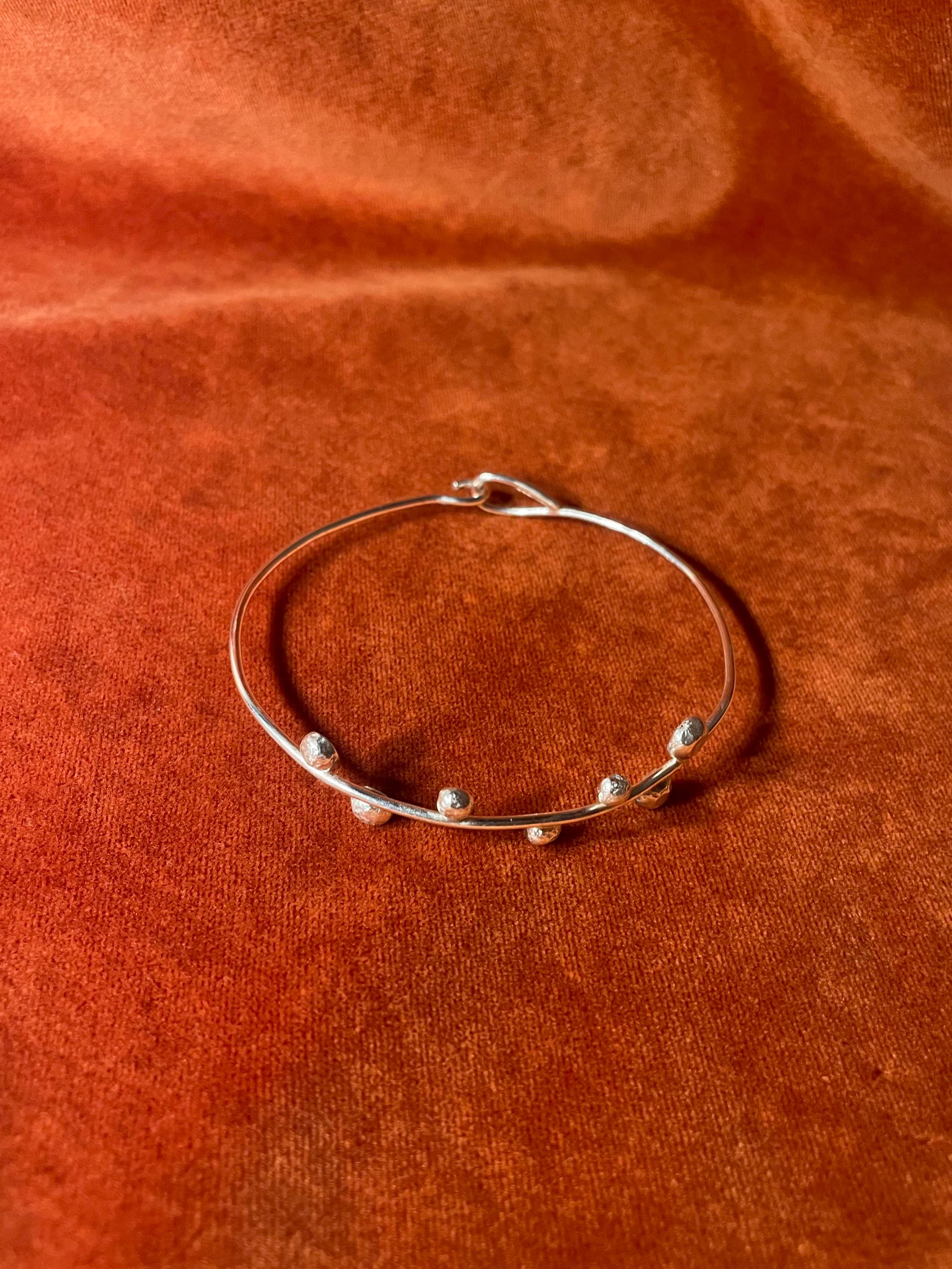 Top down view of a silver bangle with a hook clasp and seven small orbs on the top of the band. Stage on an orange velvet background.
