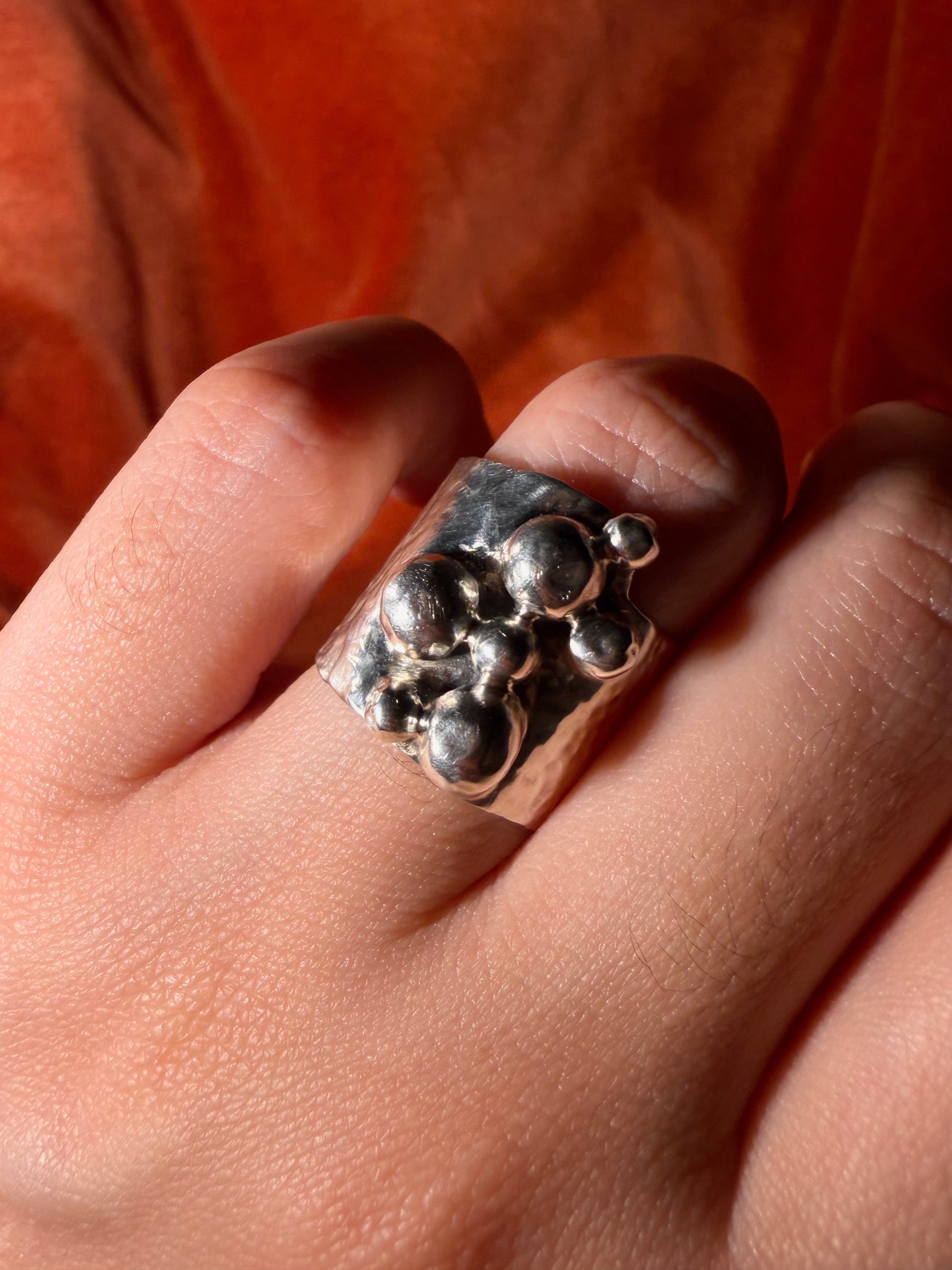 Close up image of a silver ring on a model's hand. The ring contains features a chunky hammered texture band. Seven varying sizes of orbs sit asymmetrically on top of the band.