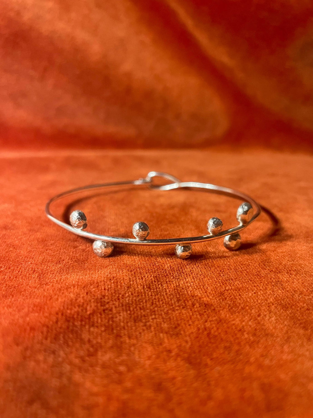 Front view of a silver bracelet with seven small orbs on the front and a closed hook clasp. Stage on an orange velvet background.