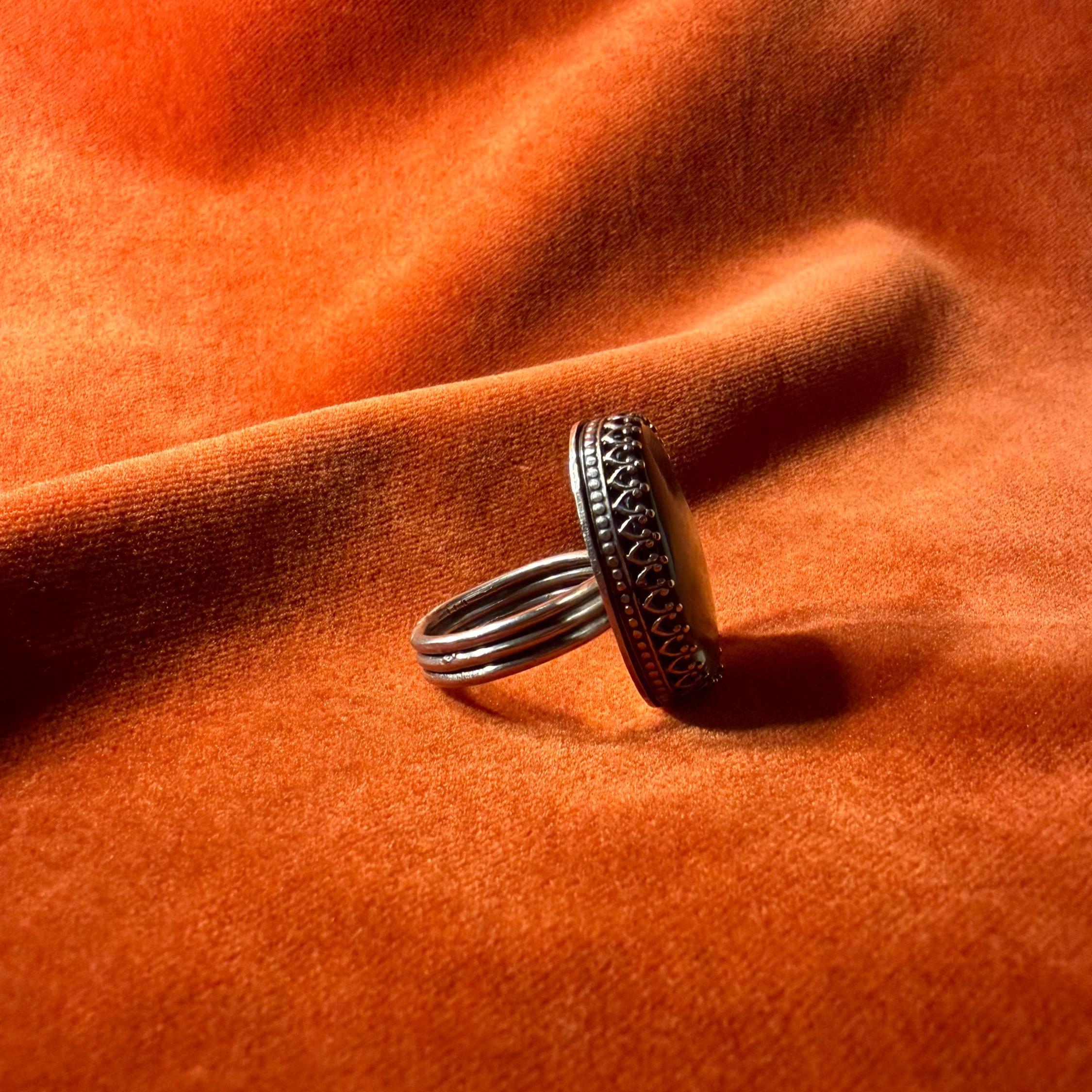 Side profile of a silver ring with a colorful abalone oval shaped stone set in an ornate bezel sitting on a medium sized band. Staged on an orange velvet background.