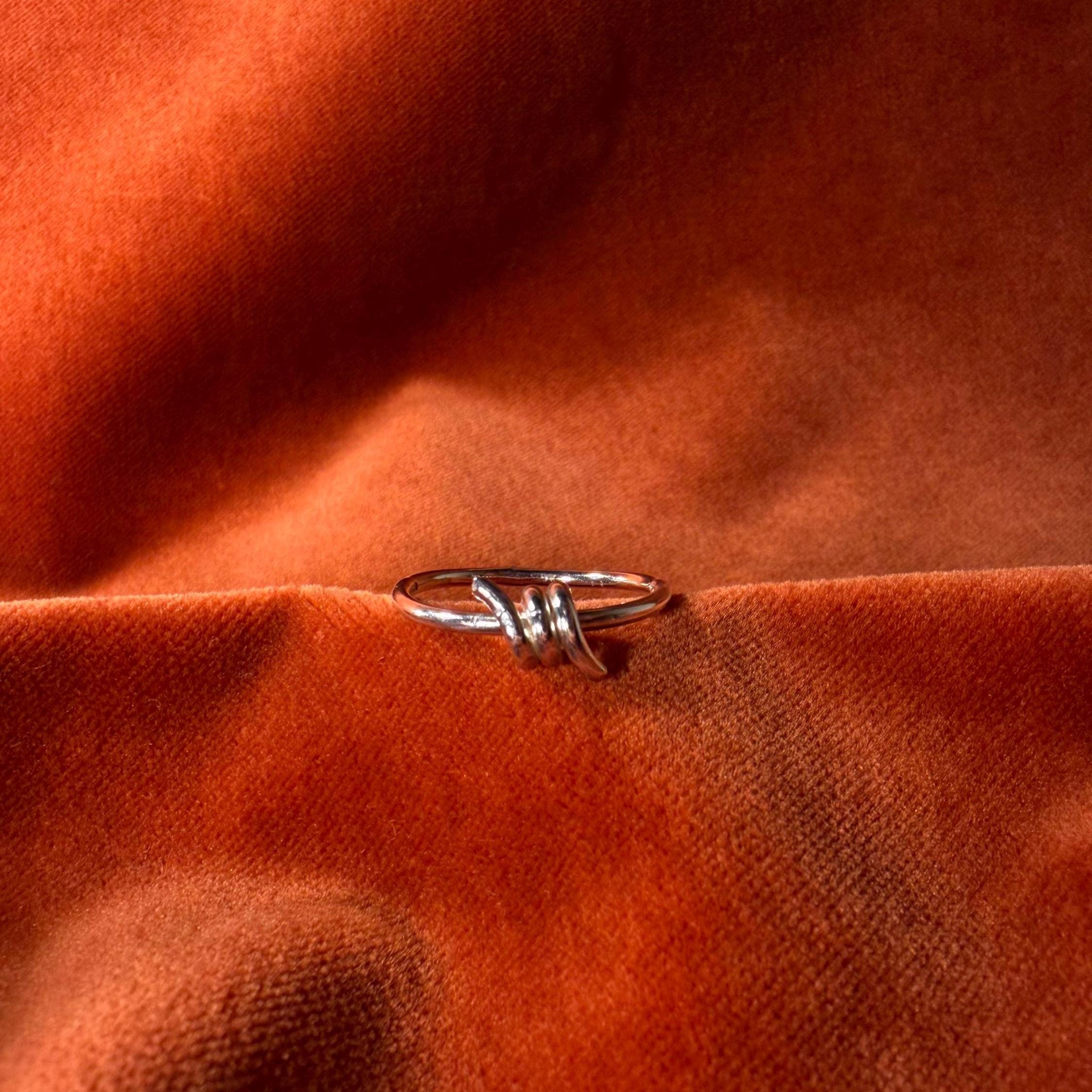 Silver ring with a design that features a barb on the top of the ring. Ring is staged on an orange velvet background.