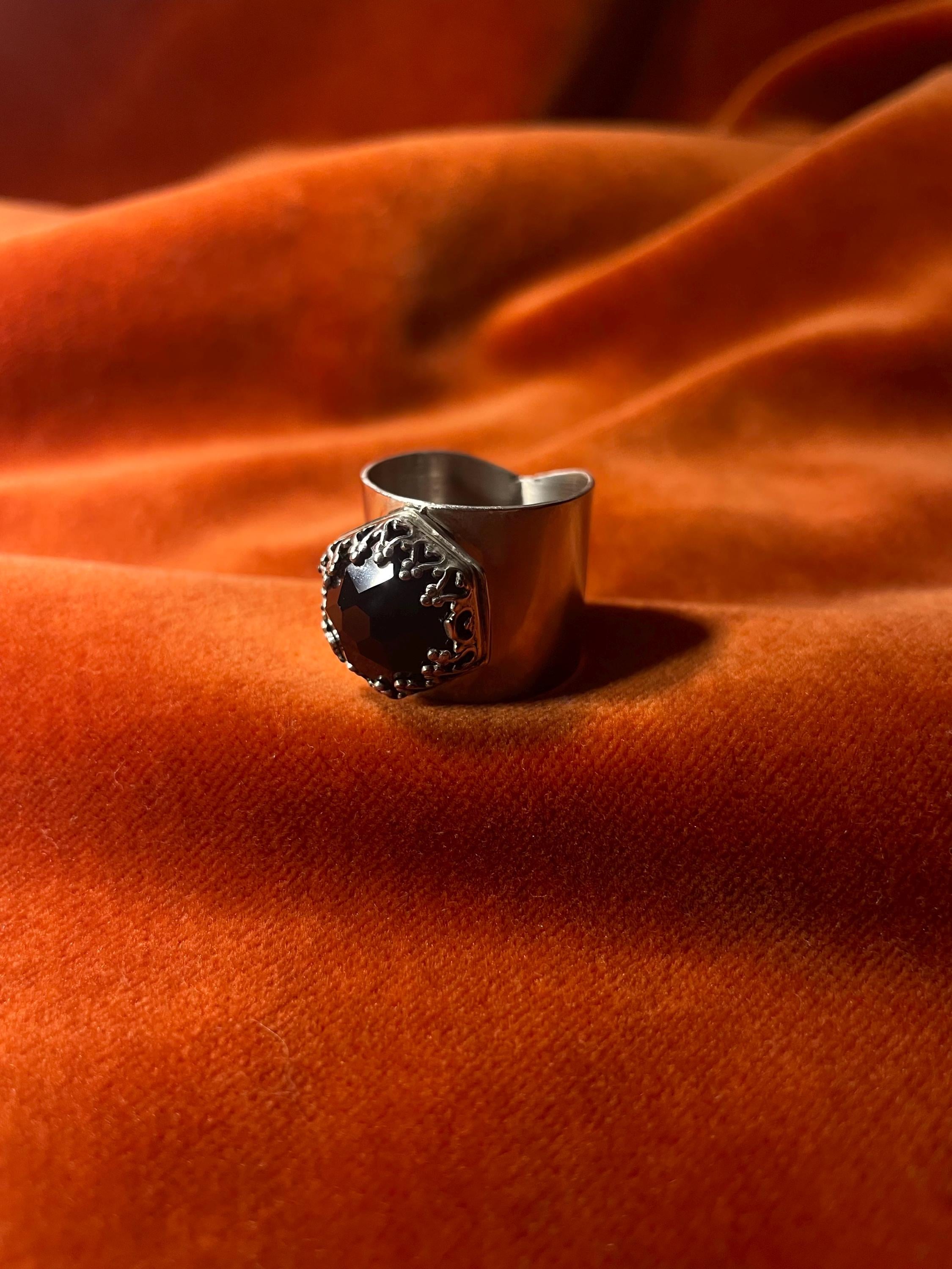 Silver ring with a black onyx rose cut stone, set on top of the large band. Ring sits against a velvet orange backdrop. 