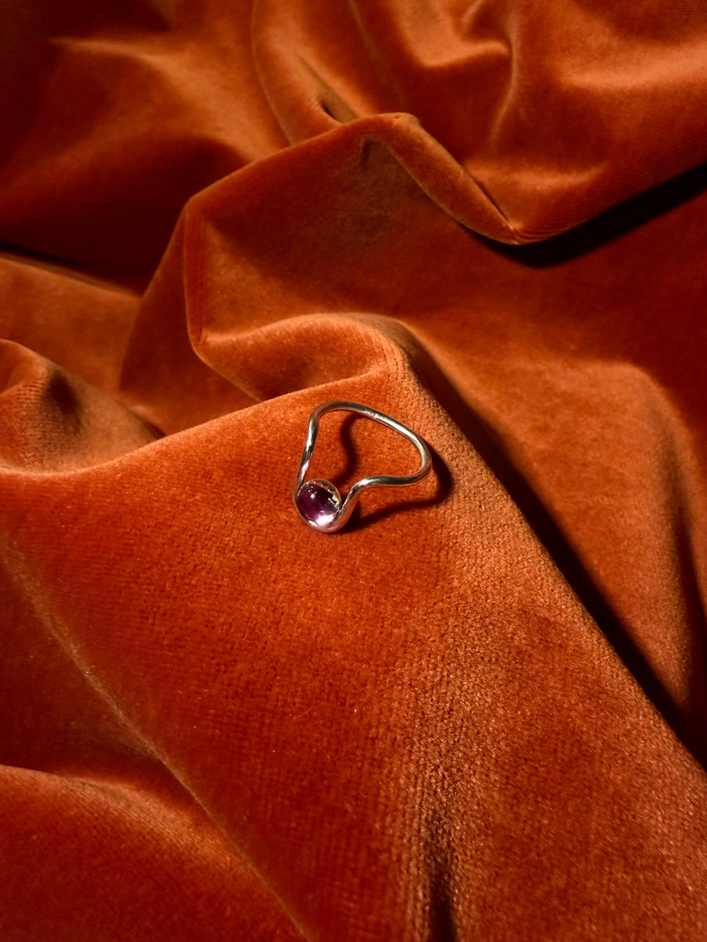 Close up image of a silver ring with bend in the ring where a serrated bezel holds a pink sapphire stone. Staged against an orange velvet background.