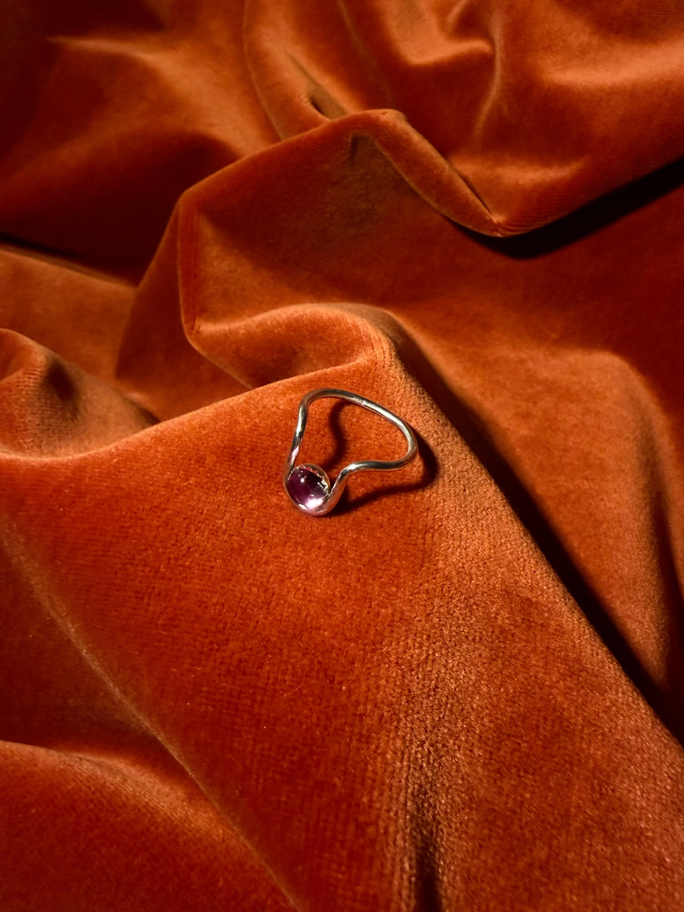 Close up image of a silver ring with bend in the ring where a serrated bezel holds a pink sapphire stone. Staged against an orange velvet background.