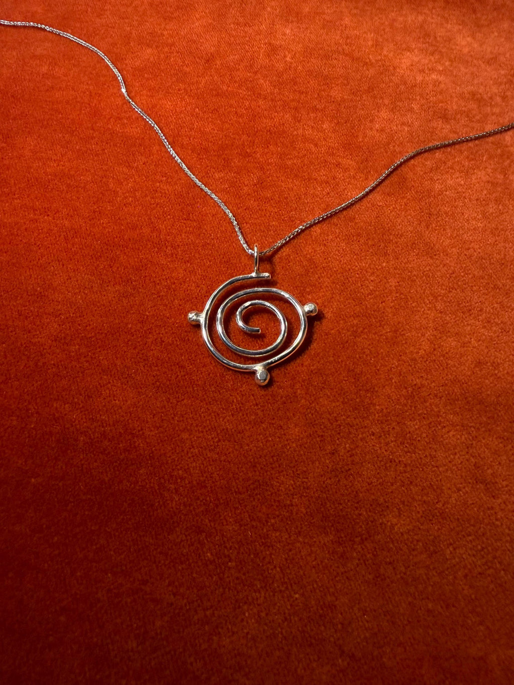 Silver spiral charm on silver chain. Three orbs surround that edge of the charm. Staged on a rust-colored background.