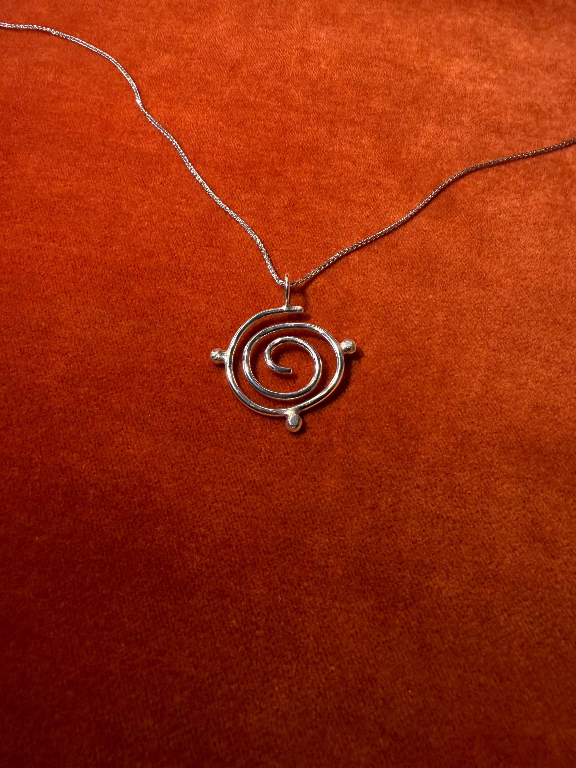 Silver spiral charm on silver chain. Three orbs surround that edge of the charm. Staged on a rust-colored background.