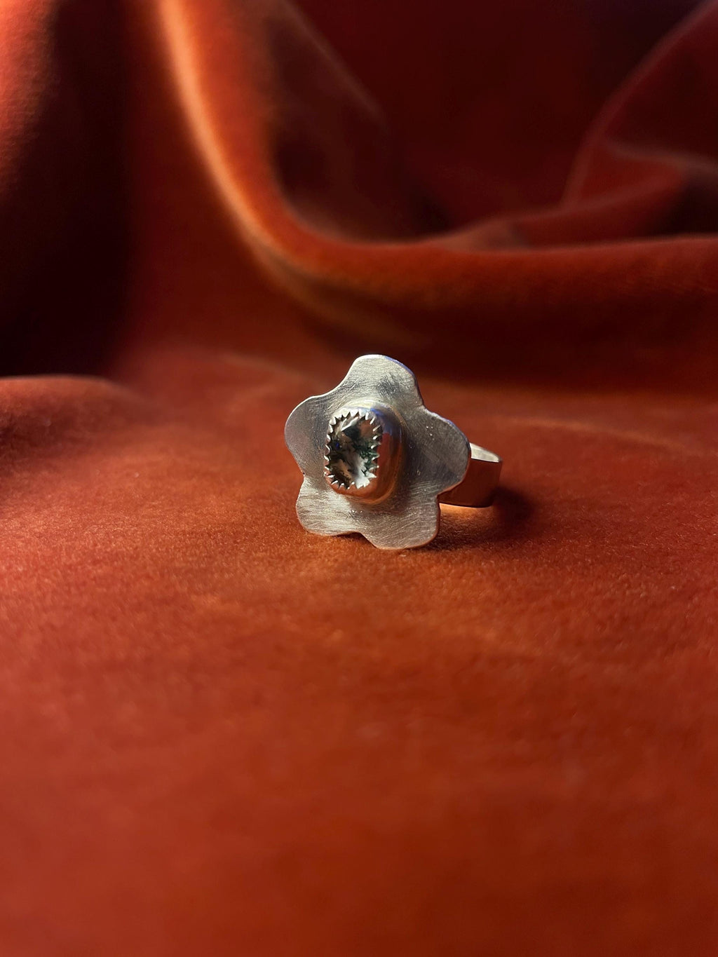 Silver ring with a flower petal design and a moss agate gemstone set in the center sitting on a small band. Staged against an orange velvet background.