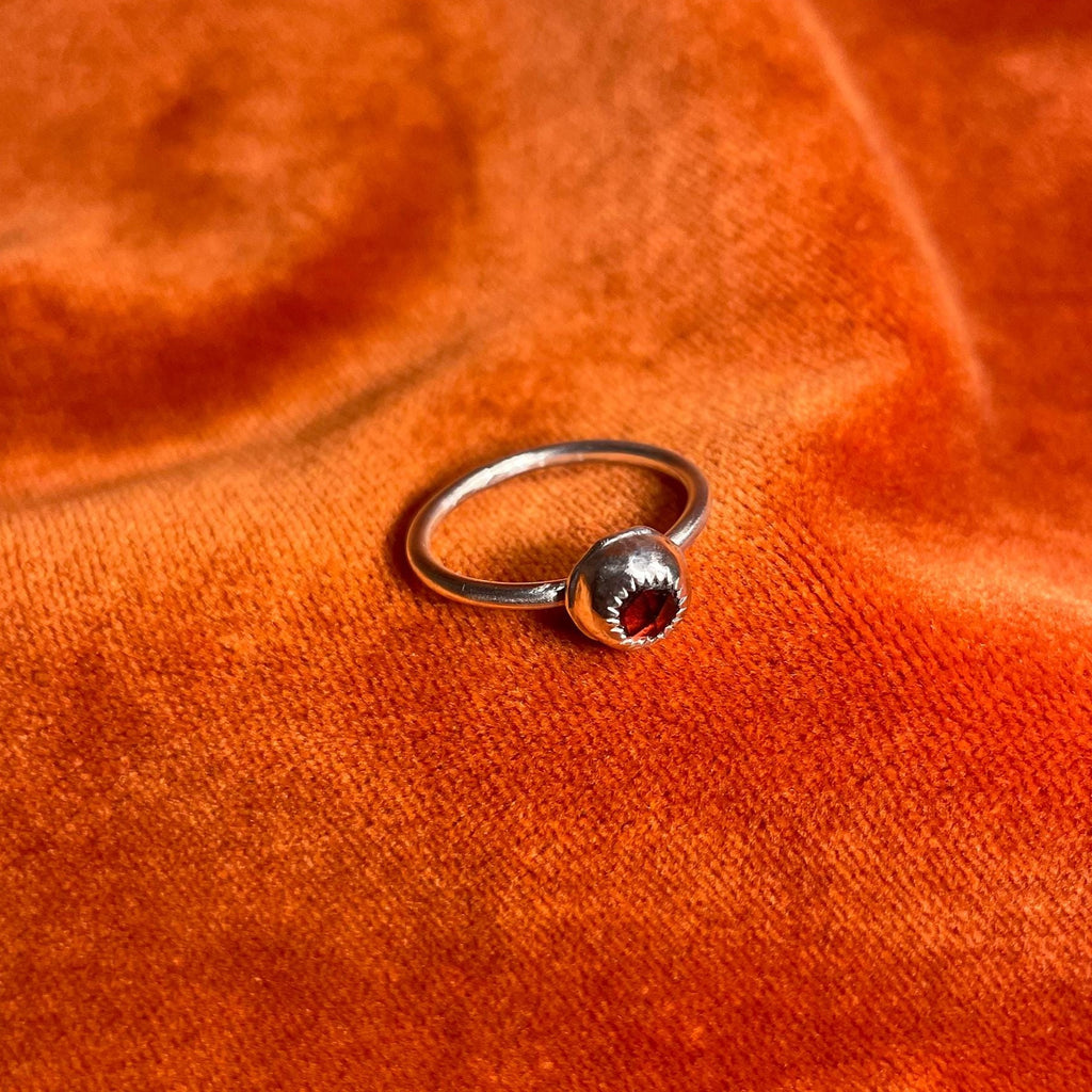 Silver ring with a serrated bezel enclosure and a rose cut garnet stone set inside sitting on a thin band. Ring is staged on an orange velvet background.