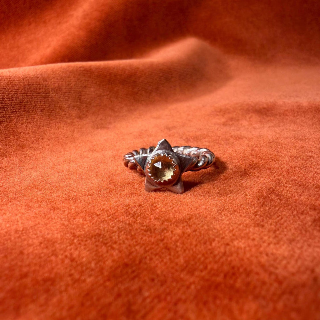 Silver ring with a rope band. A star with a golden citrine stone set in the center of the star. Staged on an orange velvet background.