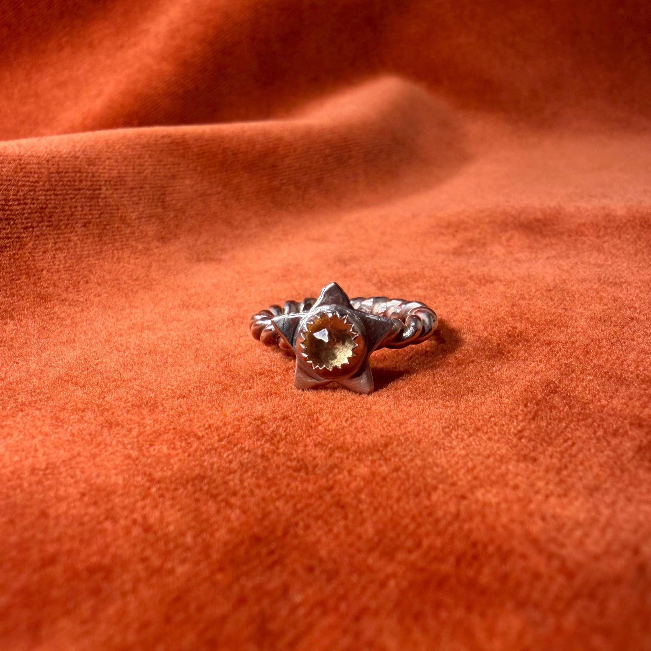 Silver ring with a rope band. A star with a golden citrine stone set in the center of the star. Staged on an orange velvet background.