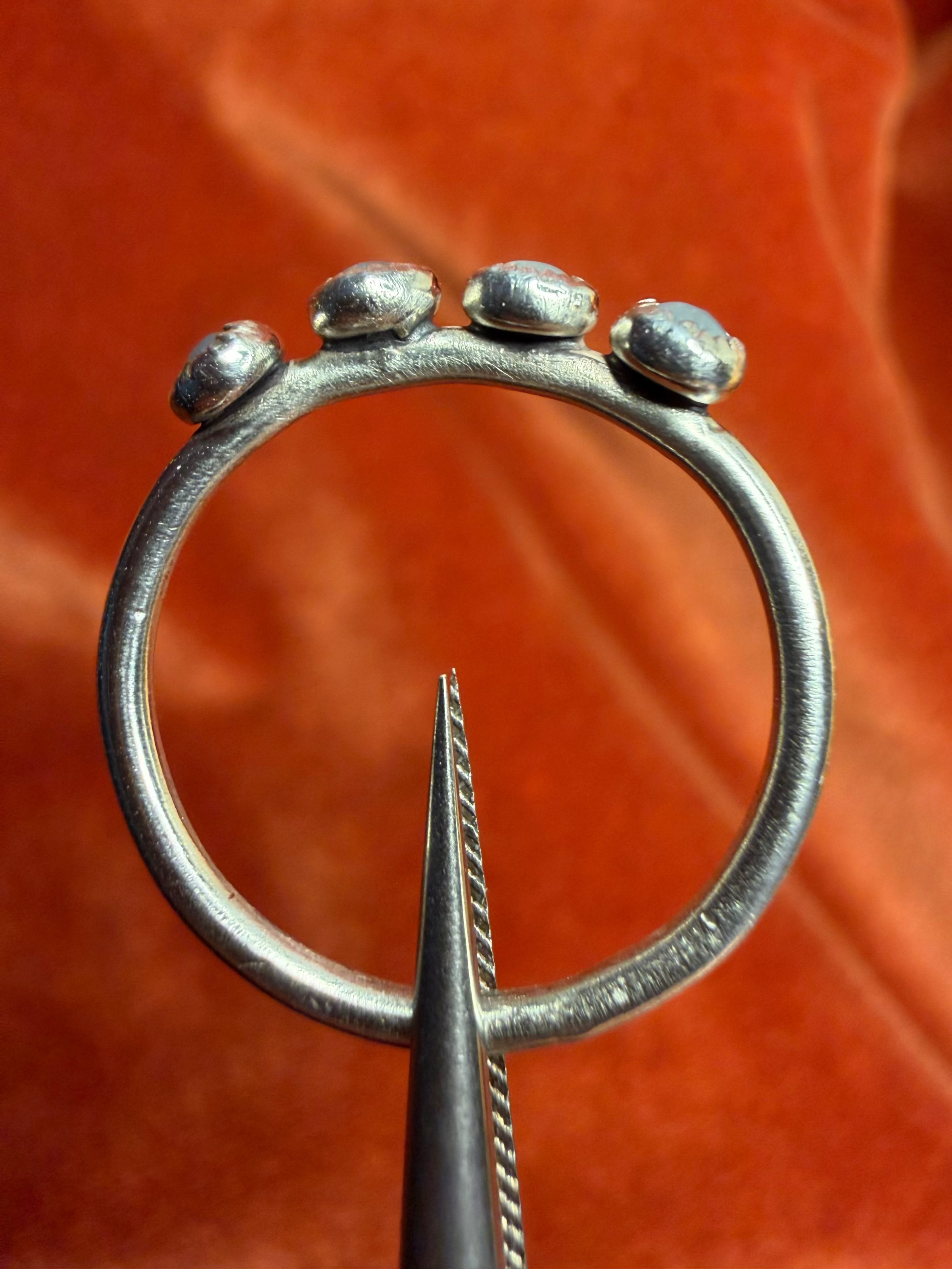 Close up image of a silver ring with four small milky blue aquamarine stones, set in serrated bezels. Laid on a small thing band. Ring is held be silver tweezers.