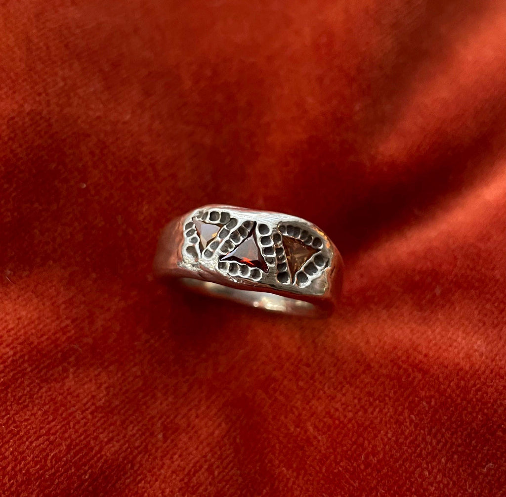 Top down view of a silver ring with three cubic zirconia triangle shaped stones. Two stones are yellow and surround one red stone. Oxidized circular impression surround the stones. 