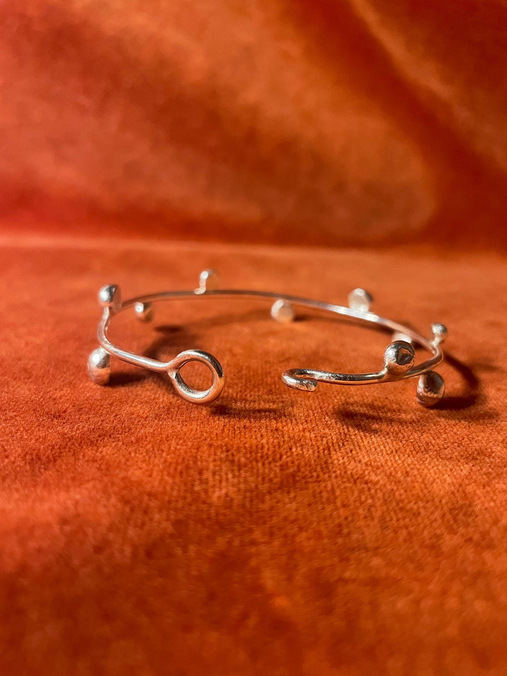 Silver bracelet showing the hook clasp opened. Bracelets features ten small orbs around the bangle. Staged on an orange velvet backdrop.