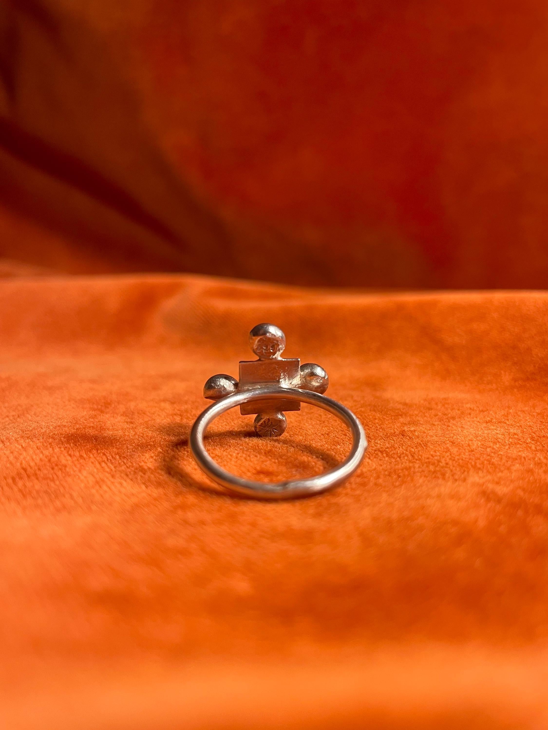 The backside of a silver ring with four orbs set around a small square shape design sitting on a thin band. Staged against an orange velvet background.