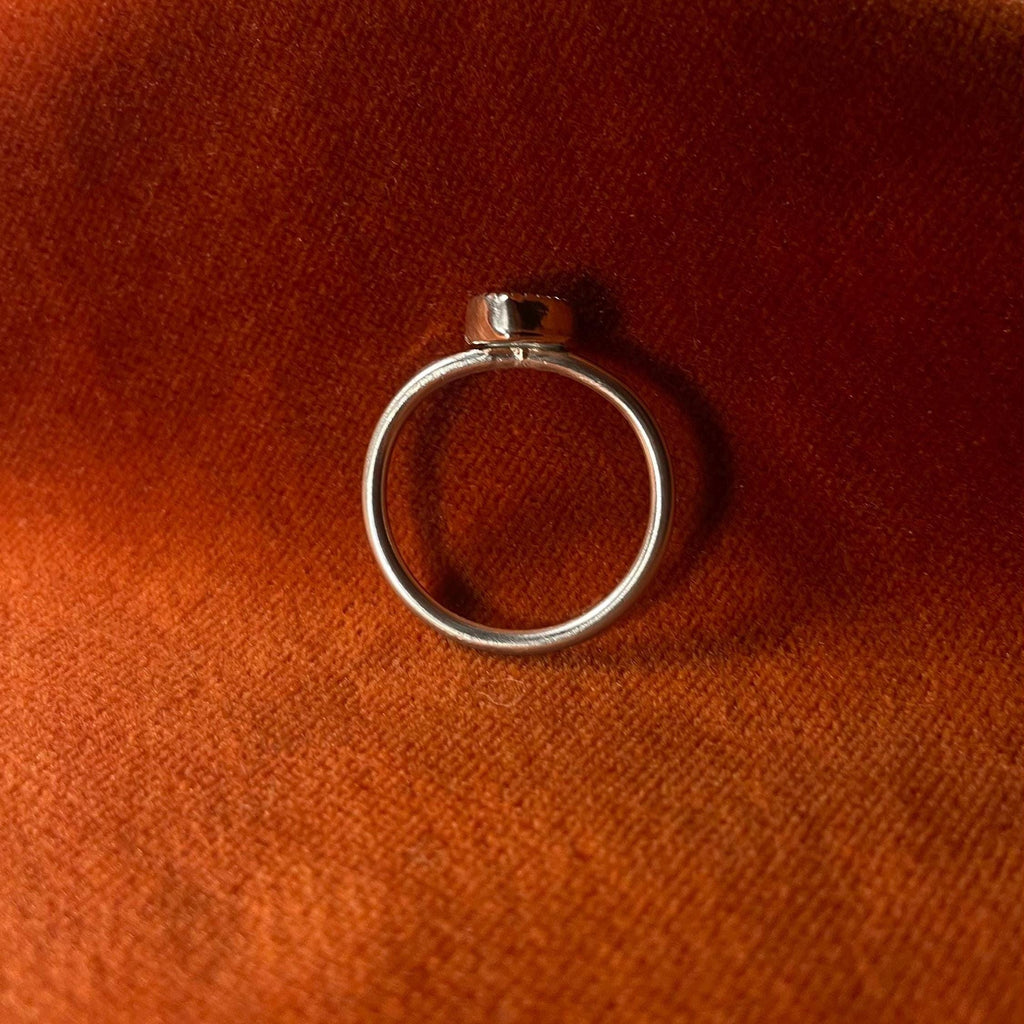 Silver ring on an orange velvet background. A pink opal heart shaped stone set in a serrated bezel sits on a thin band.