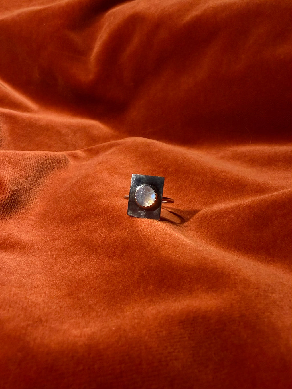 Image of a silver ring with a round rainbow white gemstone set on a square piece of silver over the band. Staged on an orange velvet background. 