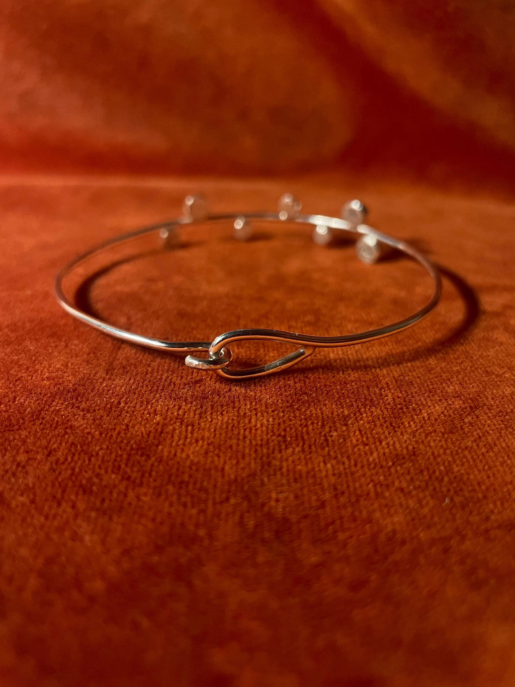 Hook clasping a silver bangle showing its closure. Seven small orbs are staggered on the top facing part of the band. Staged on an orange velvet background.