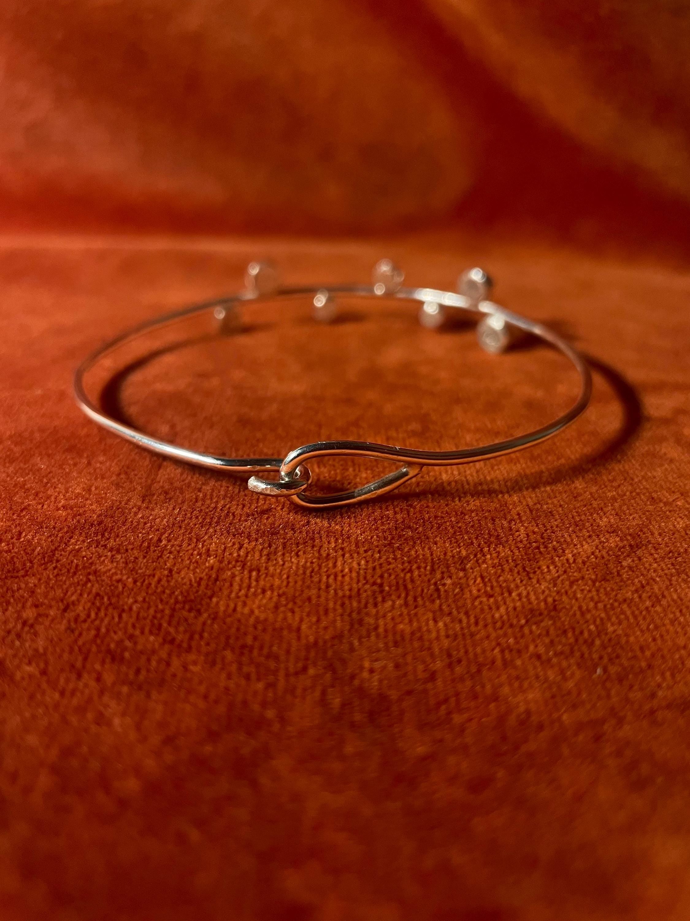 Hook clasping a silver bangle showing its closure. Seven small orbs are staggered on the top facing part of the band. Staged on an orange velvet background.