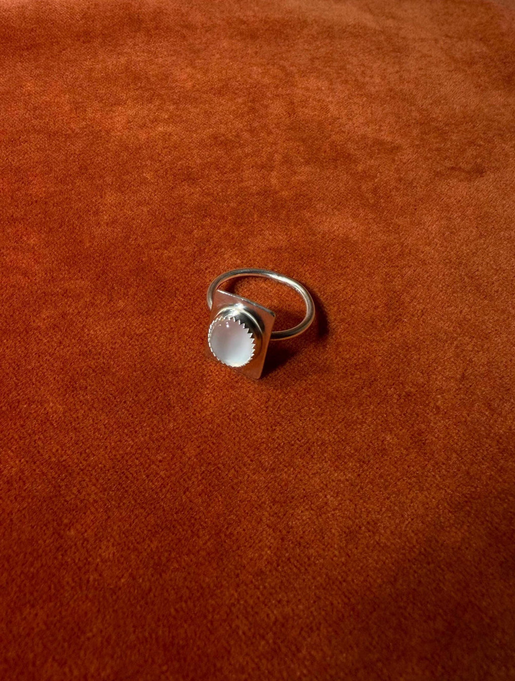 Silver ring with a serrated bezel with a white moonstone set on a rectangular fixture attached to the band of the ring. Staged on an orange velvet background.
