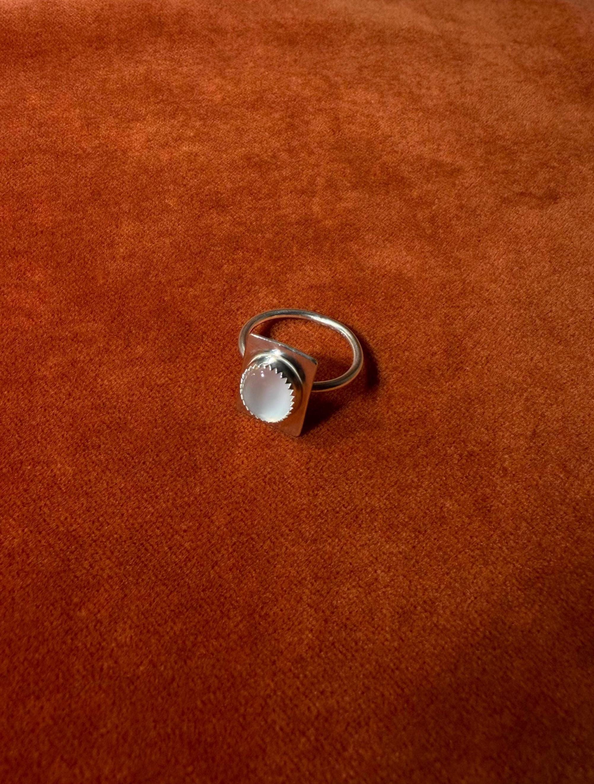 Silver ring with a serrated bezel with a white moonstone set on a rectangular fixture attached to the band of the ring. Staged on an orange velvet background.