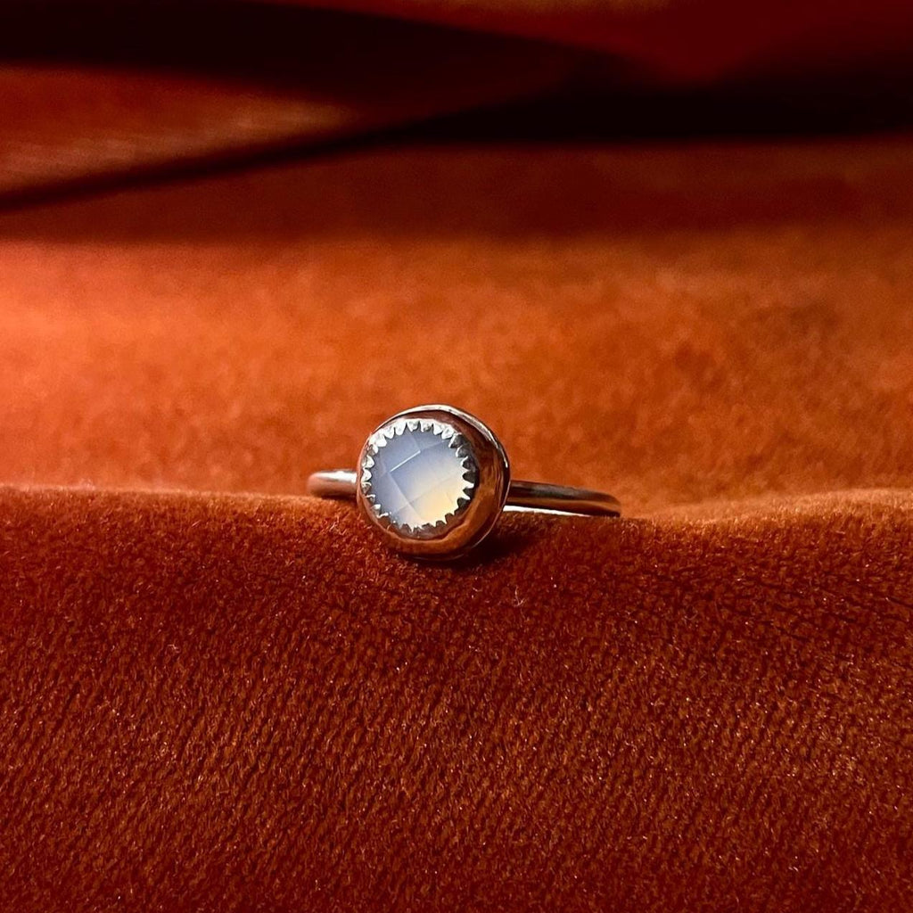 Silver ring with a sky blue chalcedony rose cut cabochon round stone set in a serrated bezel on a thin band. Set against an orange velvet background.
