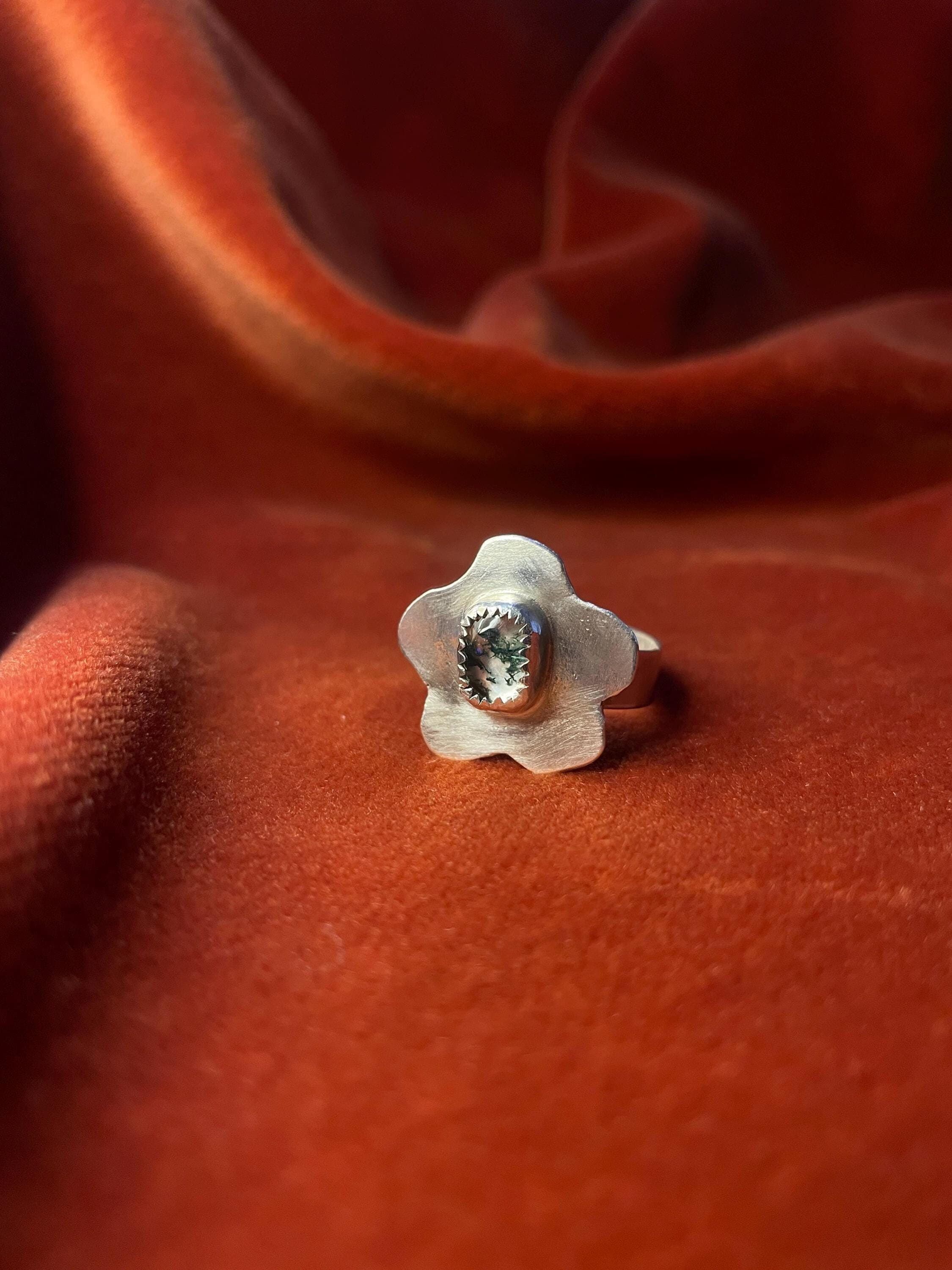 Silver ring with a flower petal design and a moss agate gemstone set in the center sitting on a small band. Staged against an orange velvet background.