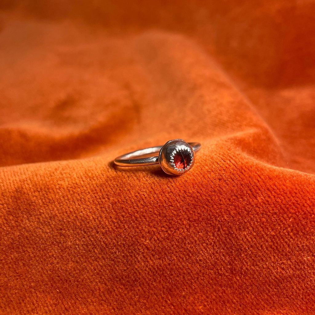 Silver ring with a serrated bezel enclosure and a rose cut garnet stone set inside sitting on a thin band. Ring is staged on an orange velvet background.