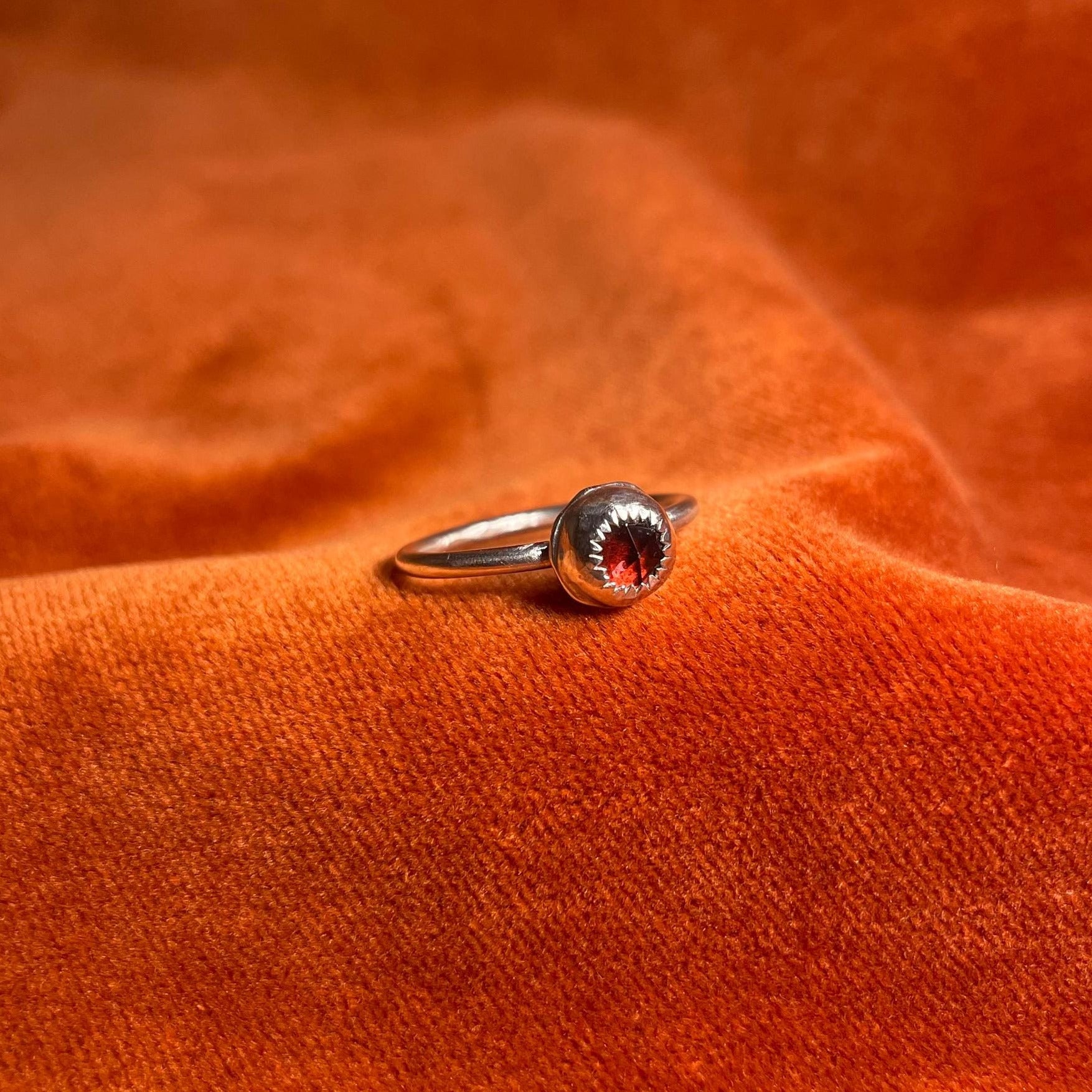 Silver ring with a serrated bezel enclosure and a rose cut garnet stone set inside sitting on a thin band. Ring is staged on an orange velvet background.