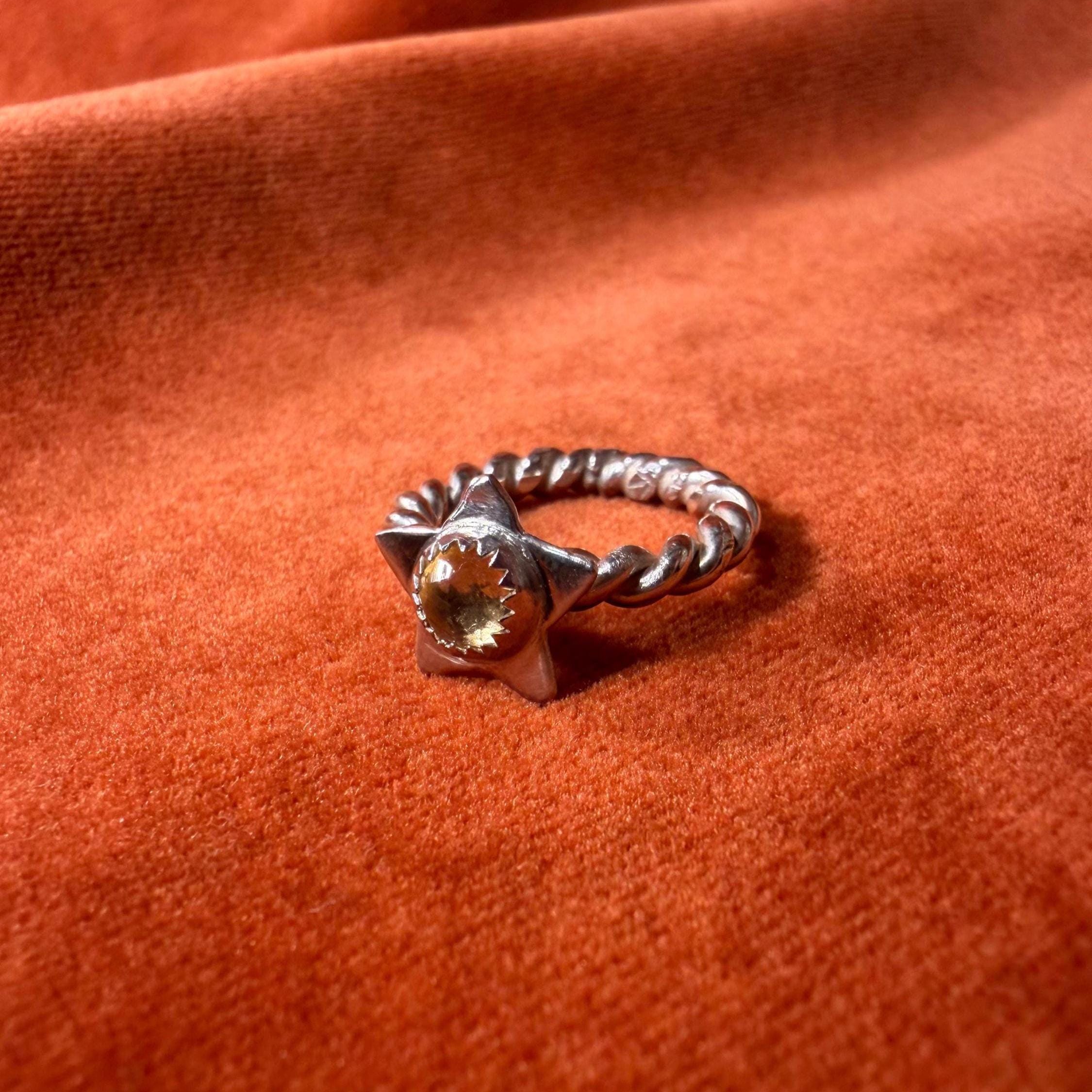 Silver ring with a rope band. A star with a golden citrine stone set in the center of the star. Staged on an orange velvet background.