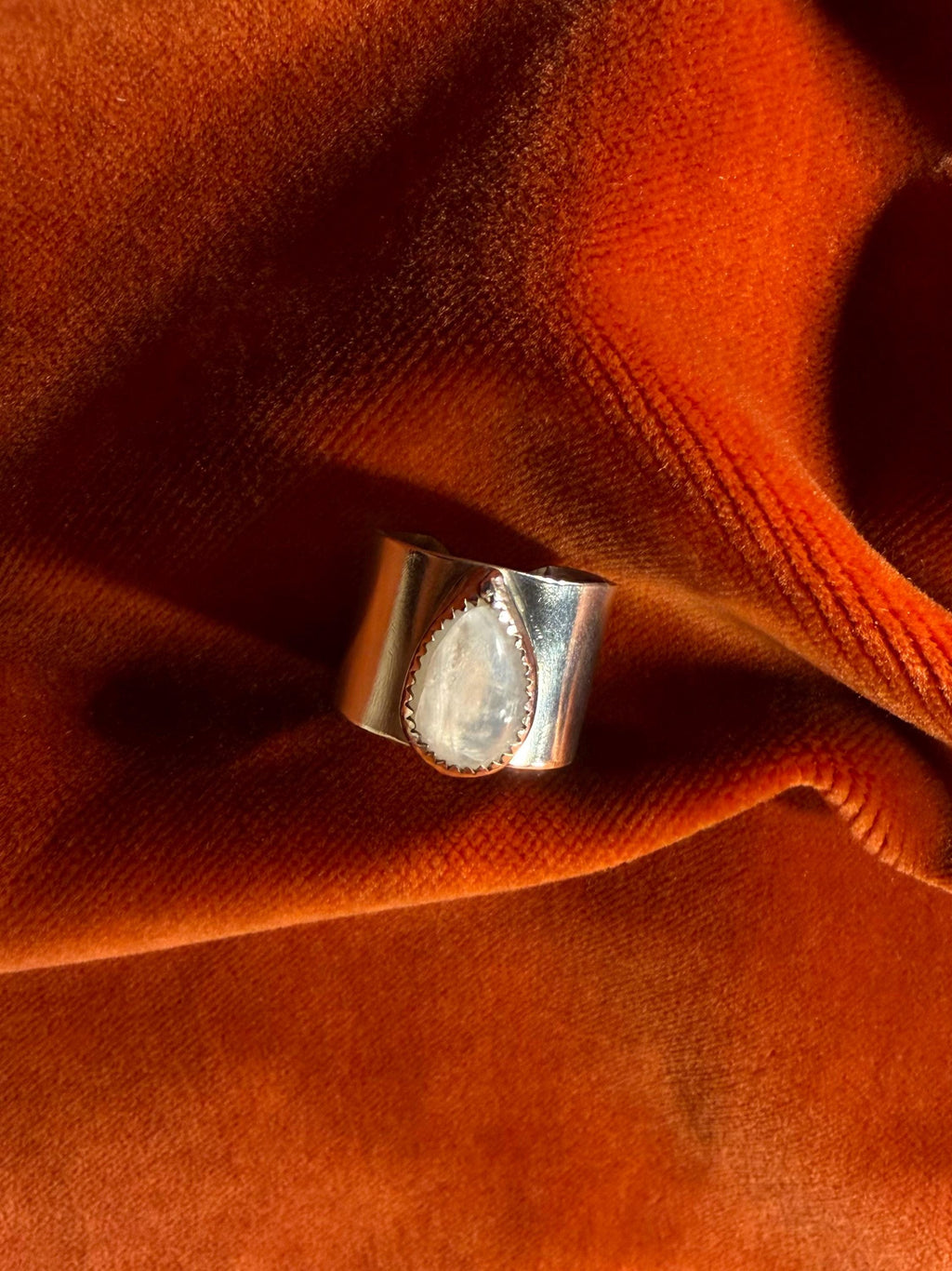 Silver ring with a teardrop shaped moonstone set on top of a chunky band against an orange velvet background.