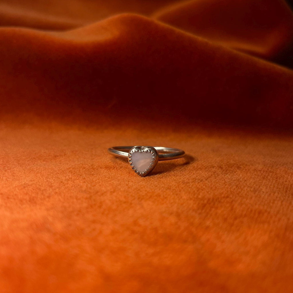 Silver ring on an orange velvet background. A pink opal heart shaped stone set in a serrated bezel sits on a thin band.