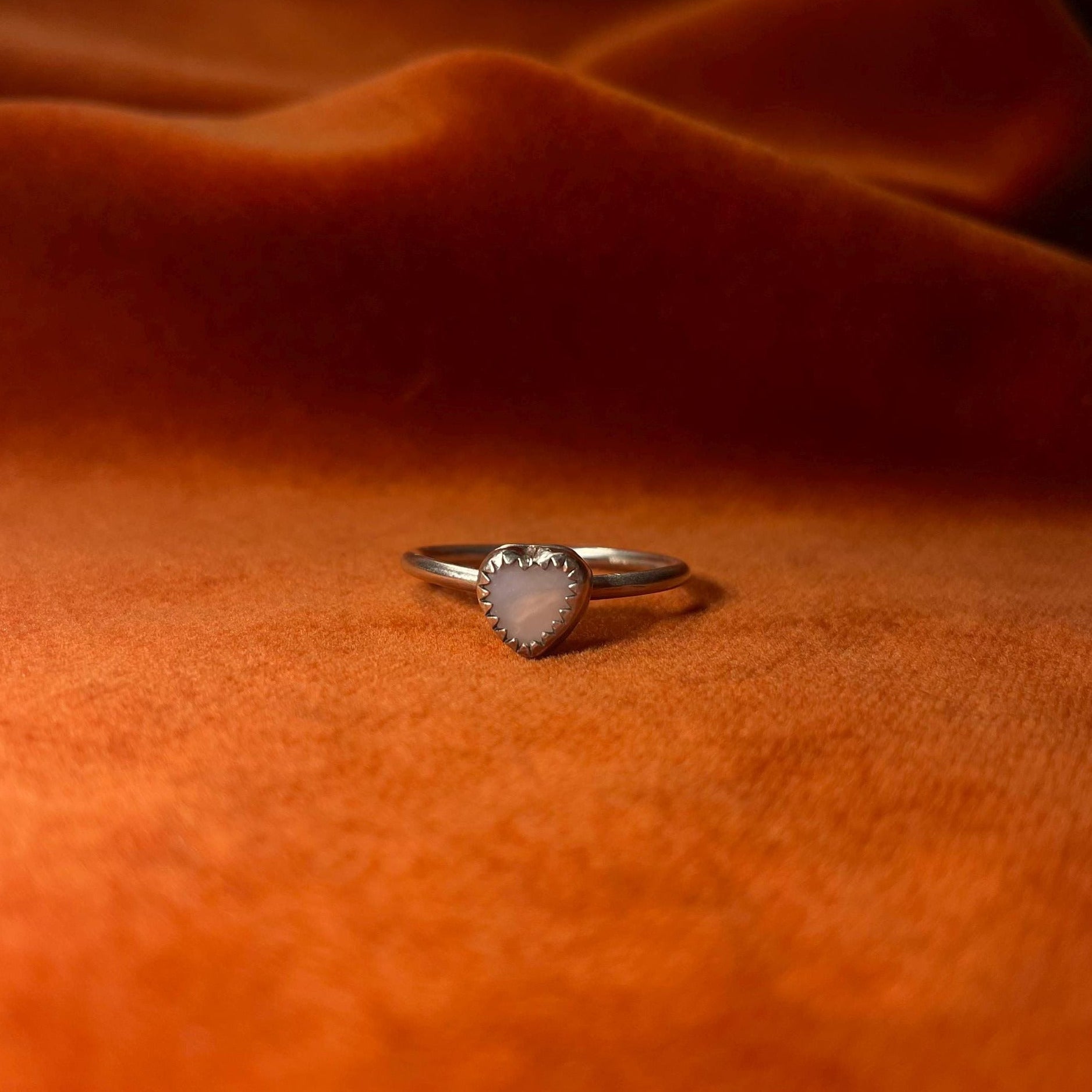 Silver ring on an orange velvet background. A pink opal heart shaped stone set in a serrated bezel sits on a thin band.