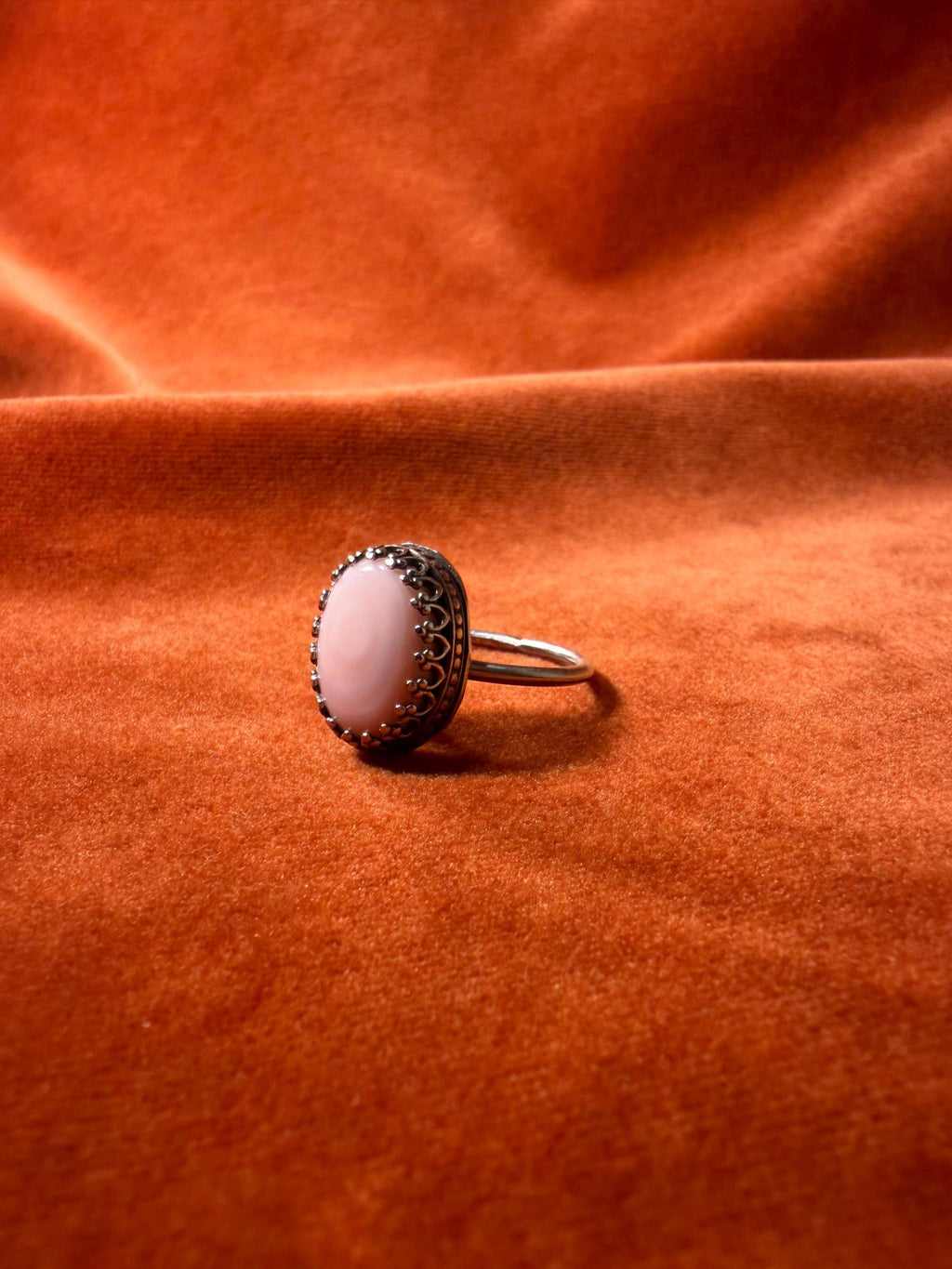 Silver ring with a pink conch stone set in an ornate bezel on a thin band. Set against an orange velvet background.