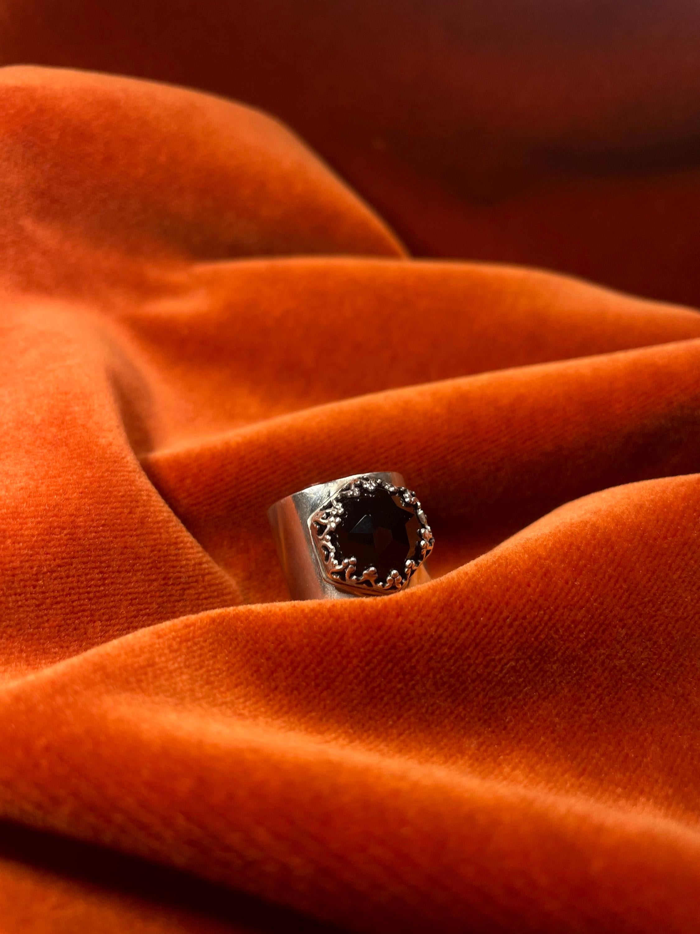 Silver ring with a black onyx rose cut stone, set on top of the large band. Ring sits against a velvet orange backdrop. 
