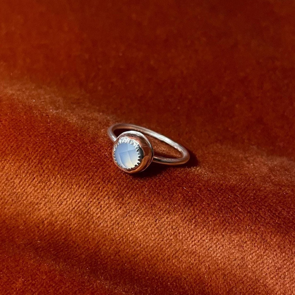 Silver ring with a sky blue chalcedony rose cut cabochon round stone set in a serrated bezel on a thin band. Set against an orange velvet background.