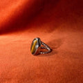 Silver ring with a roped band and a large tiger's eye stone set in a serrated bezel. Staged against an orange velvet background. 