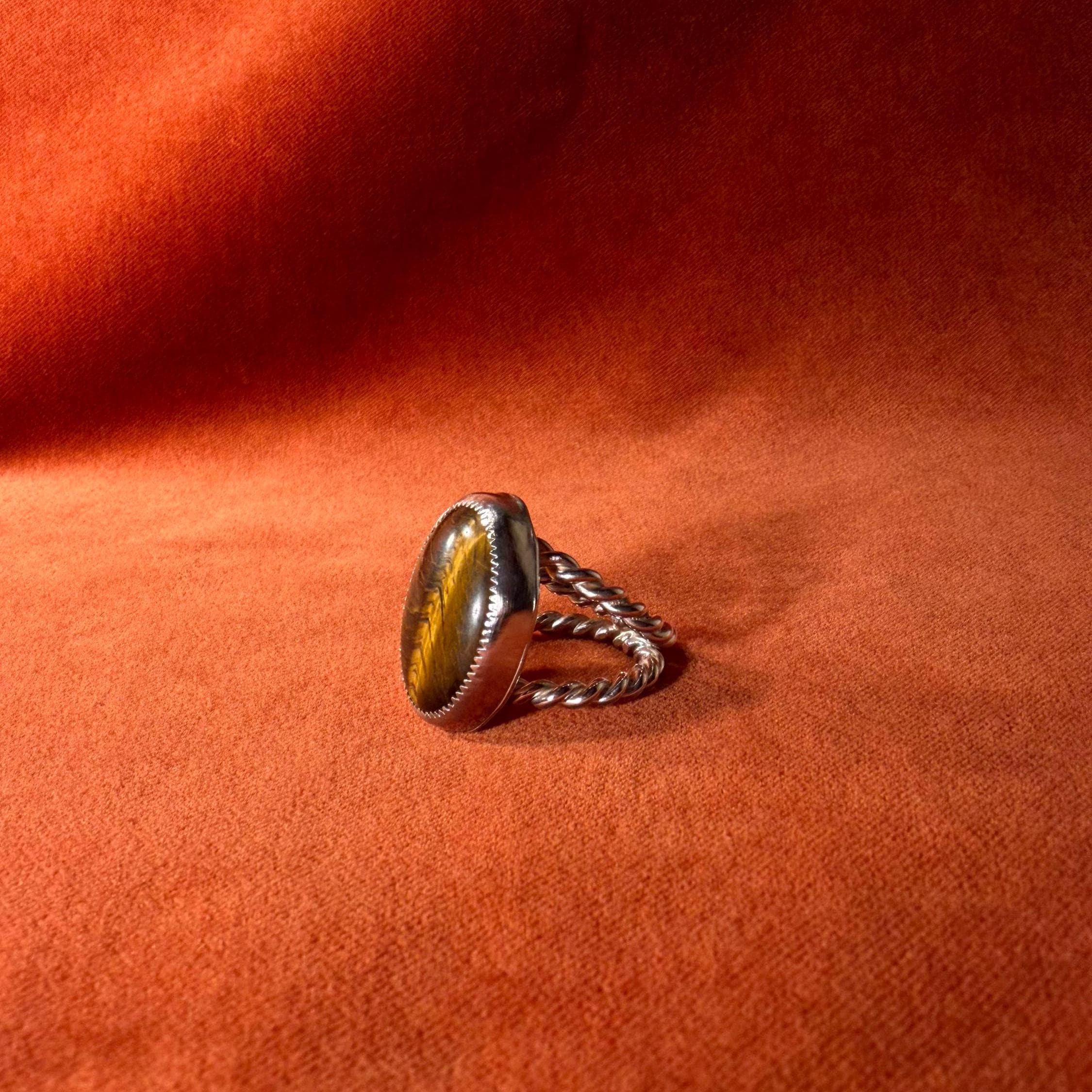 Silver ring with a roped band and a large tiger's eye stone set in a serrated bezel. Staged against an orange velvet background. 