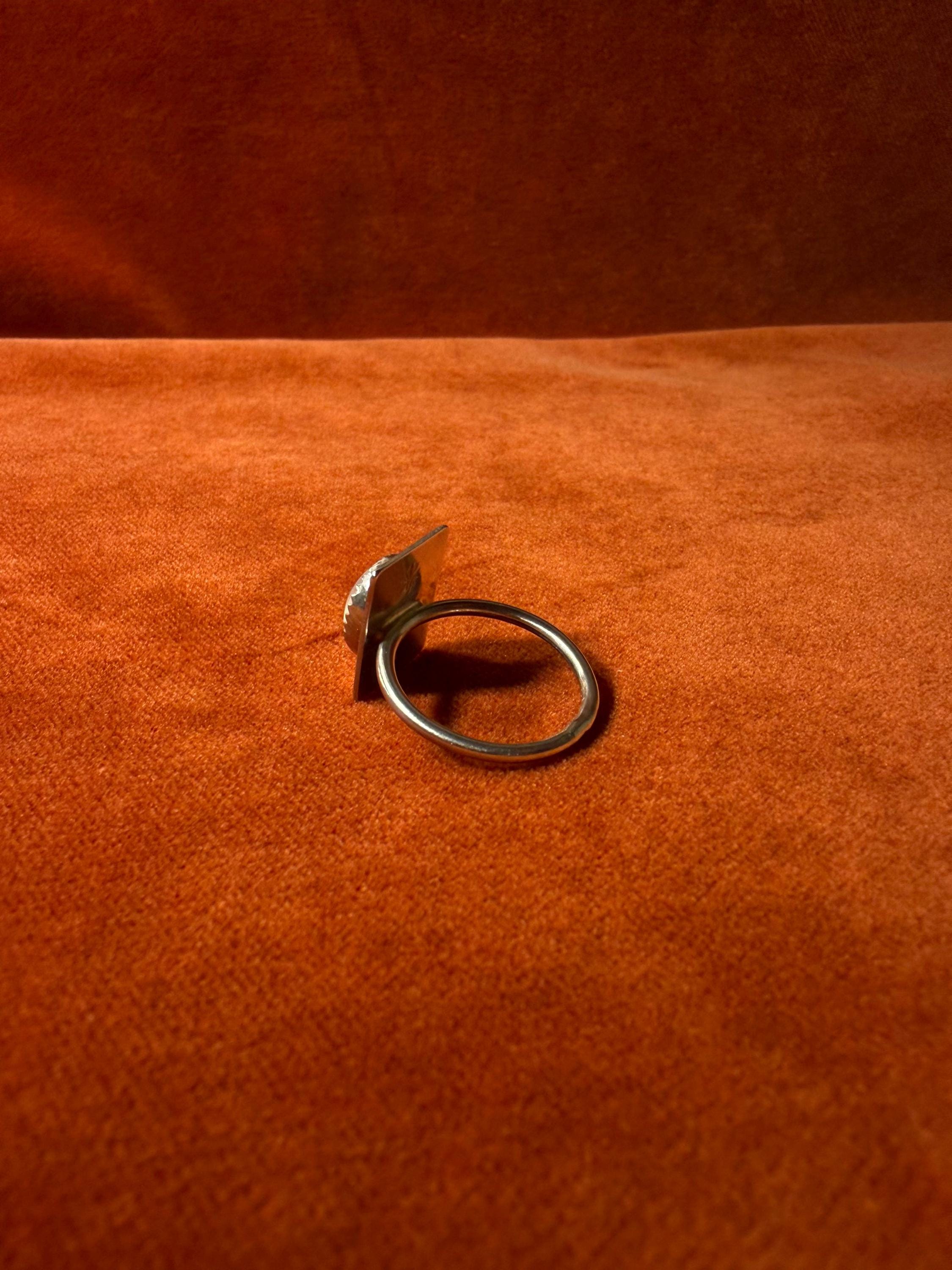 Silver ring with a serrated bezel with a white moonstone set on a rectangular fixture attached to the band of the ring. Staged on an orange velvet background.