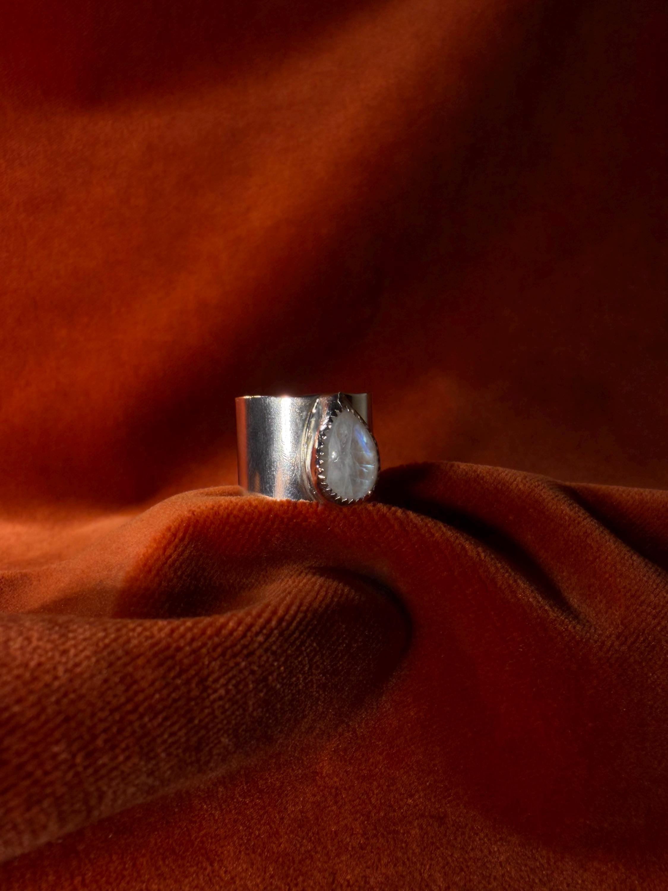 Silver ring with a teardrop shaped moonstone set on top of a chunky band against an orange velvet background.