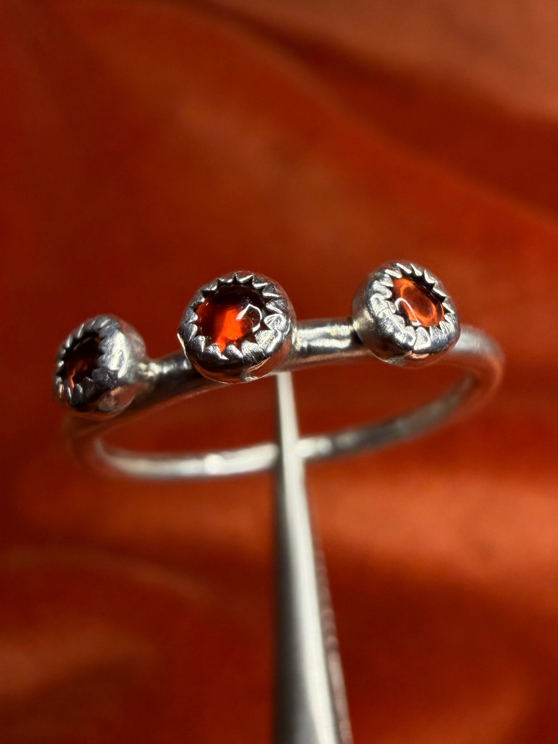 Close up image of a silver ring with three small garnet stones set in a sterling silver serrated bezel on small thin band. Staged on a orange velvet background, held by silver tweezers. 