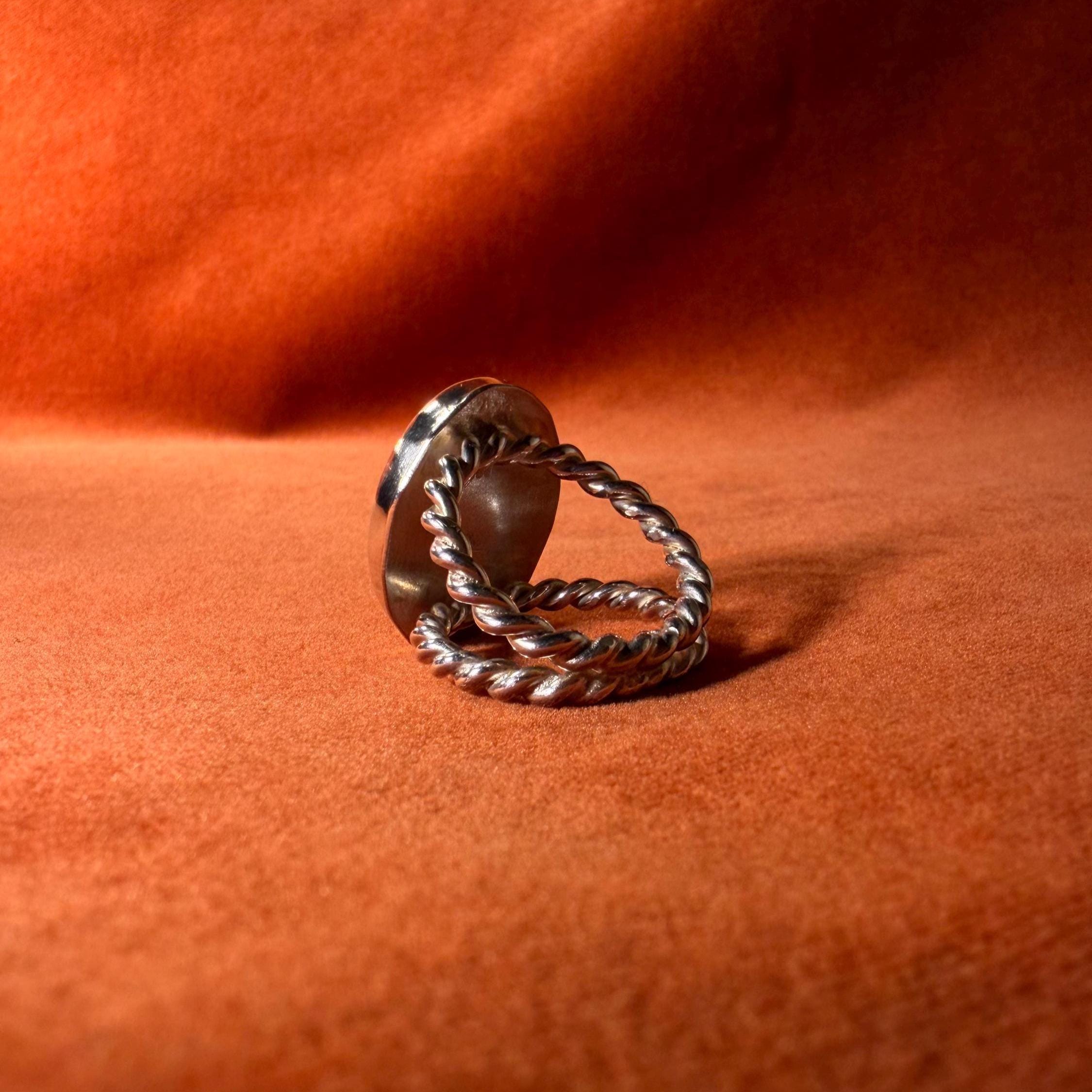 Backside of a silver ring with a roped band. Staged against an orange velvet background. 