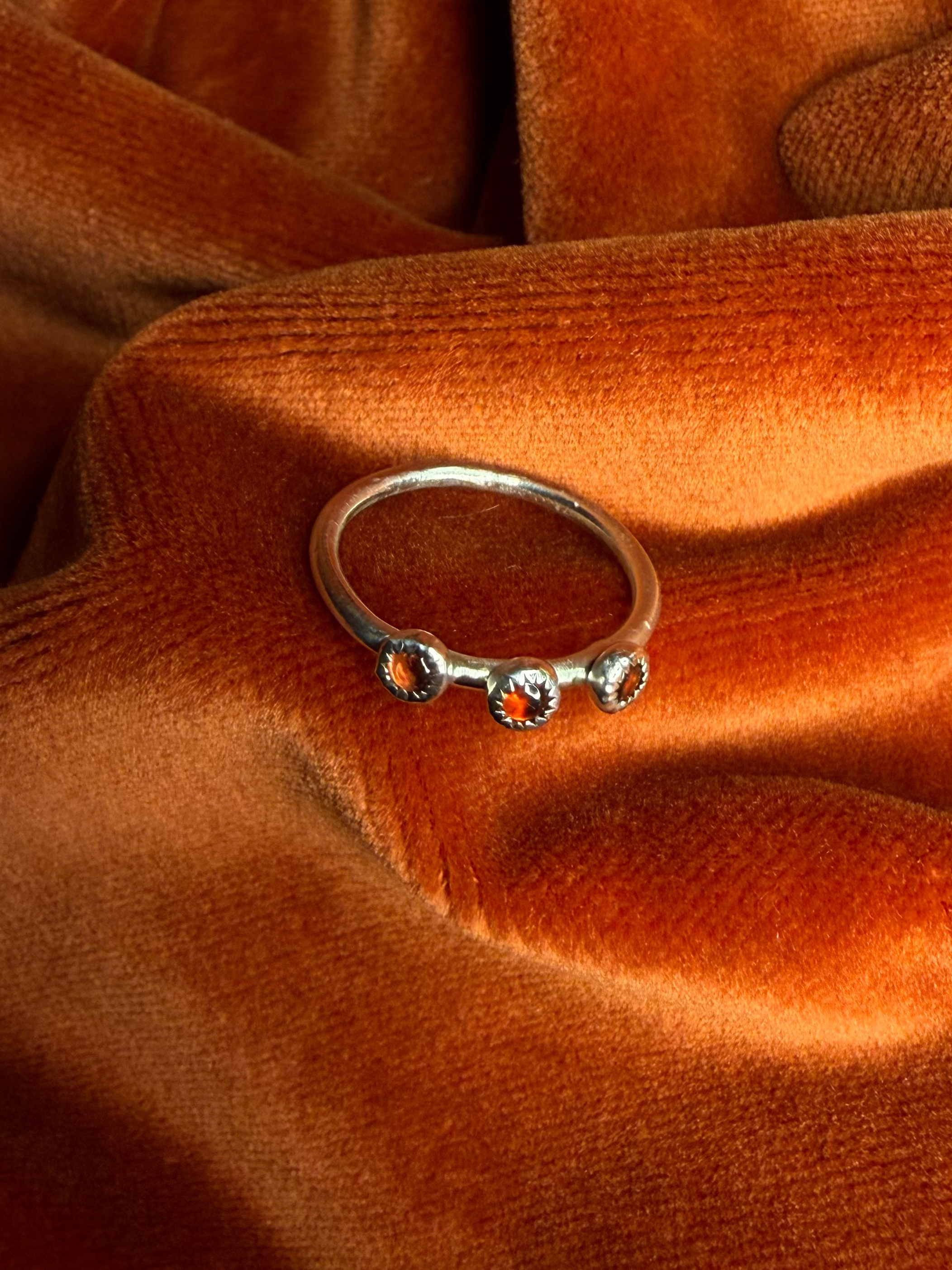Silver ring with three small garnet stones set in a sterling silver serrated bezel on small thin band. Staged on a orange velvet background.