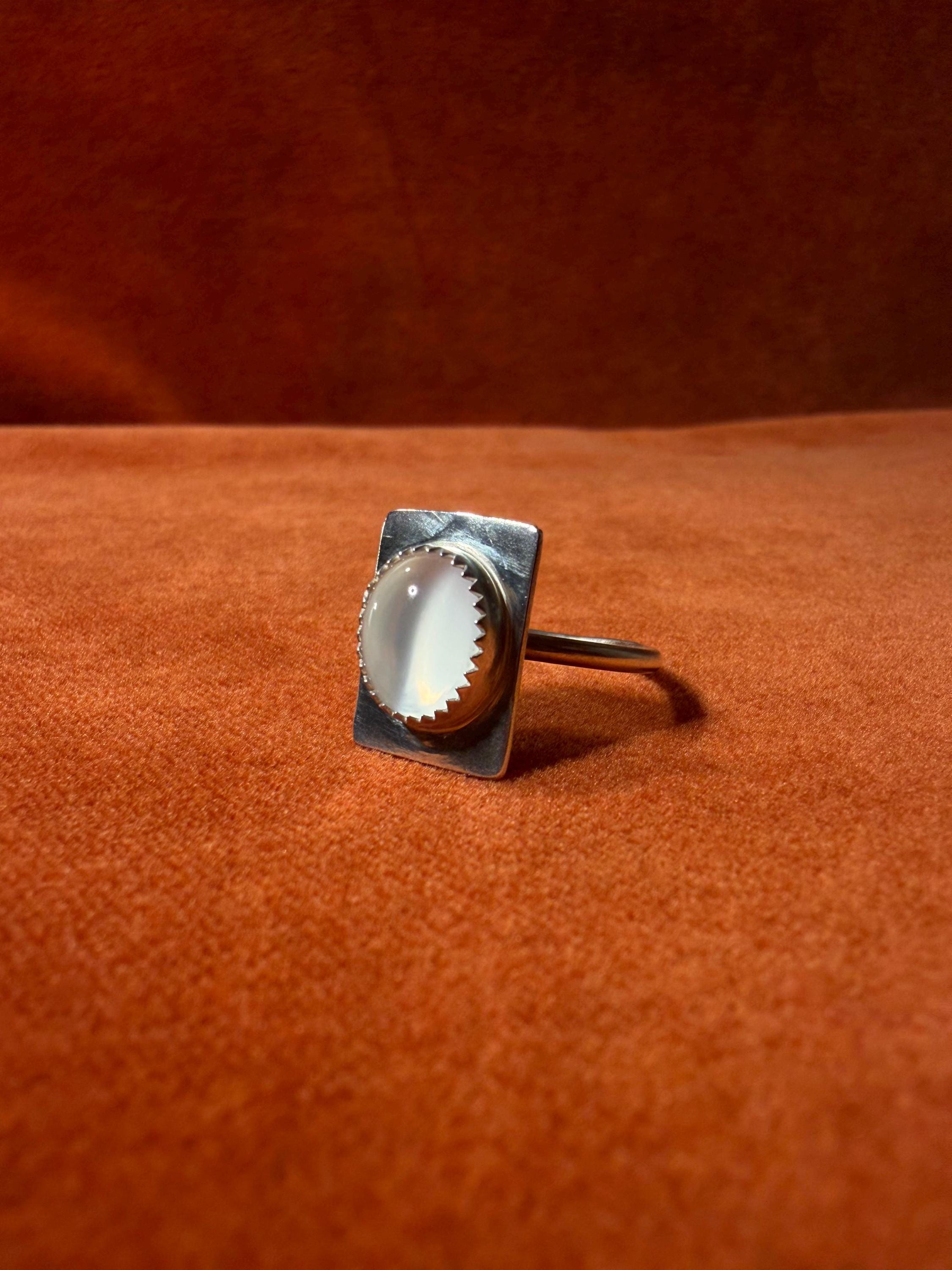 Silver ring with a serrated bezel with a white moonstone set on a rectangular fixture attached to the band of the ring. Staged on an orange velvet background.