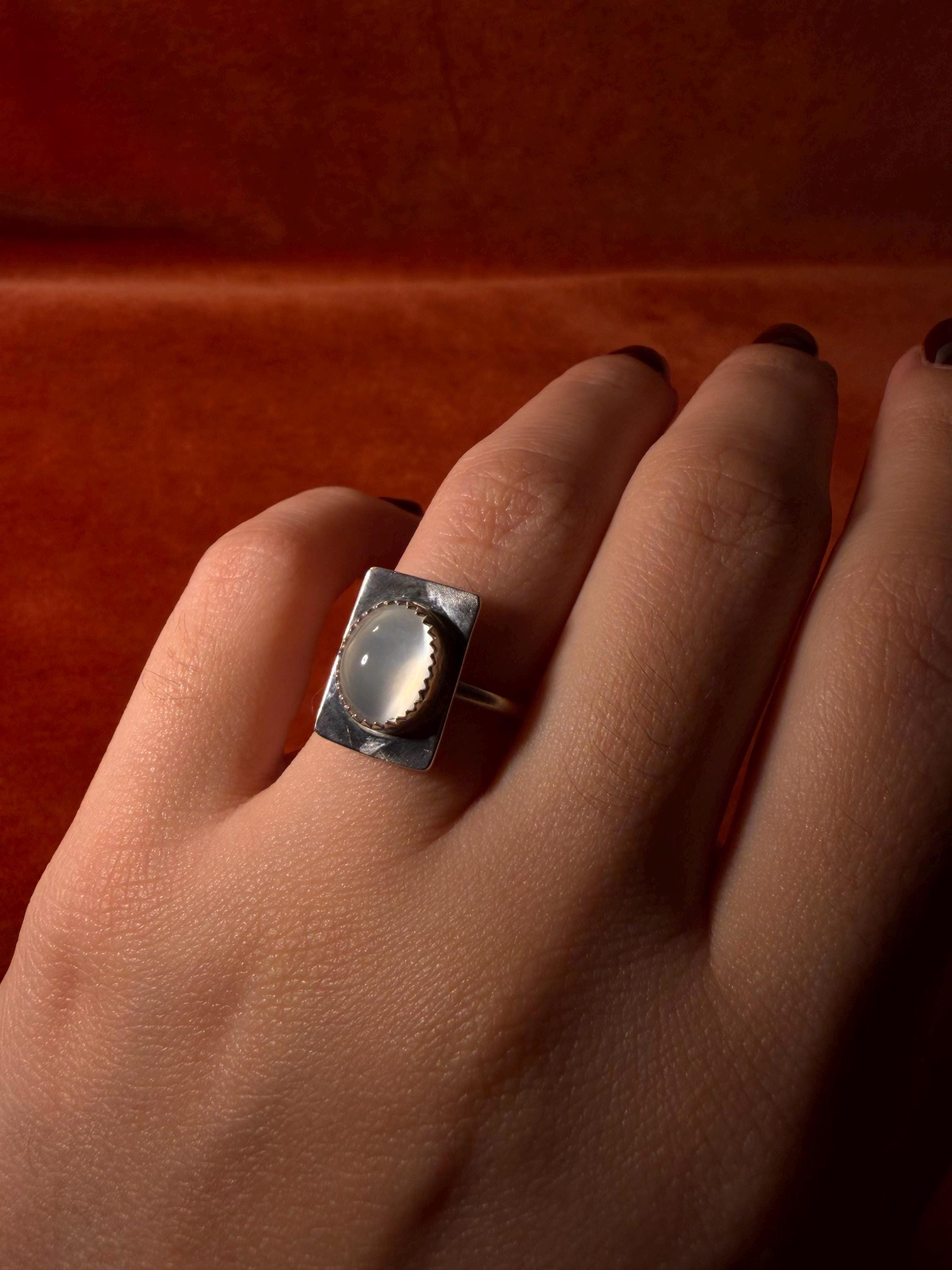 Hand modeling a silver ring with a serrated bezel with a white moonstone set on a rectangular fixture attached to the band of the ring. Staged on an orange velvet background.