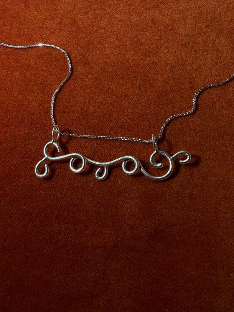 Silver chain with a silver charm designed in the shape of a wind swirls. Staged on an orange velvet background. 
